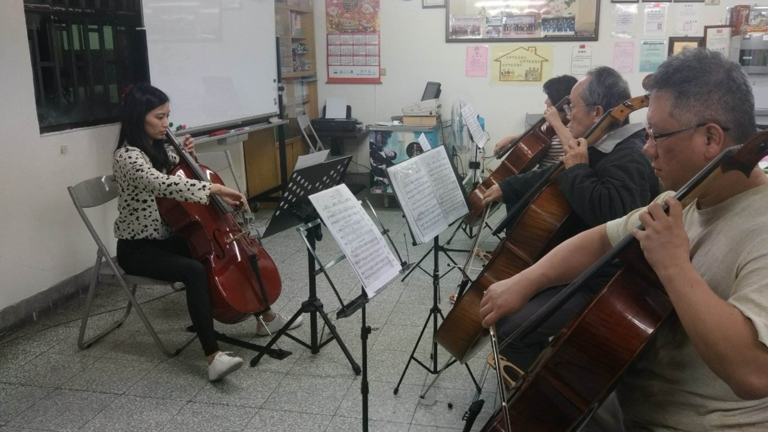 A young woman playing the cello in a classroom with four other adults, who are also playing string instruments, reading sheet music on music stands.