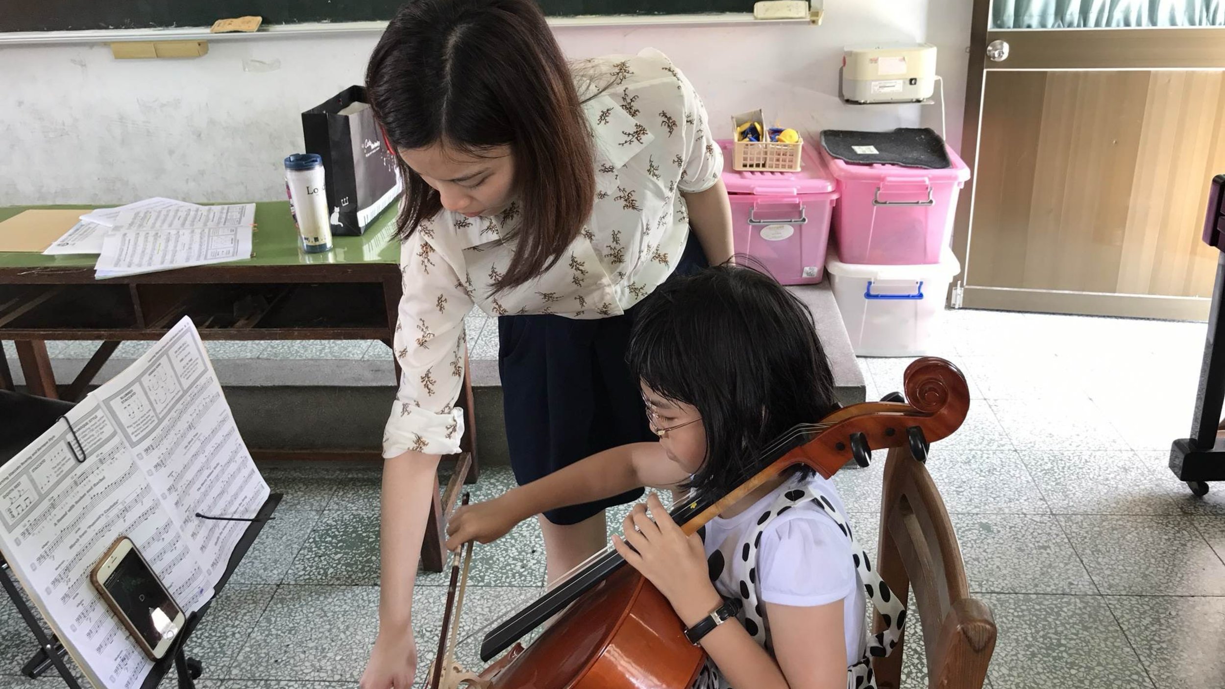 A woman helping a young girl play the cello in a classroom setting, with music sheets on a stand in front of them.