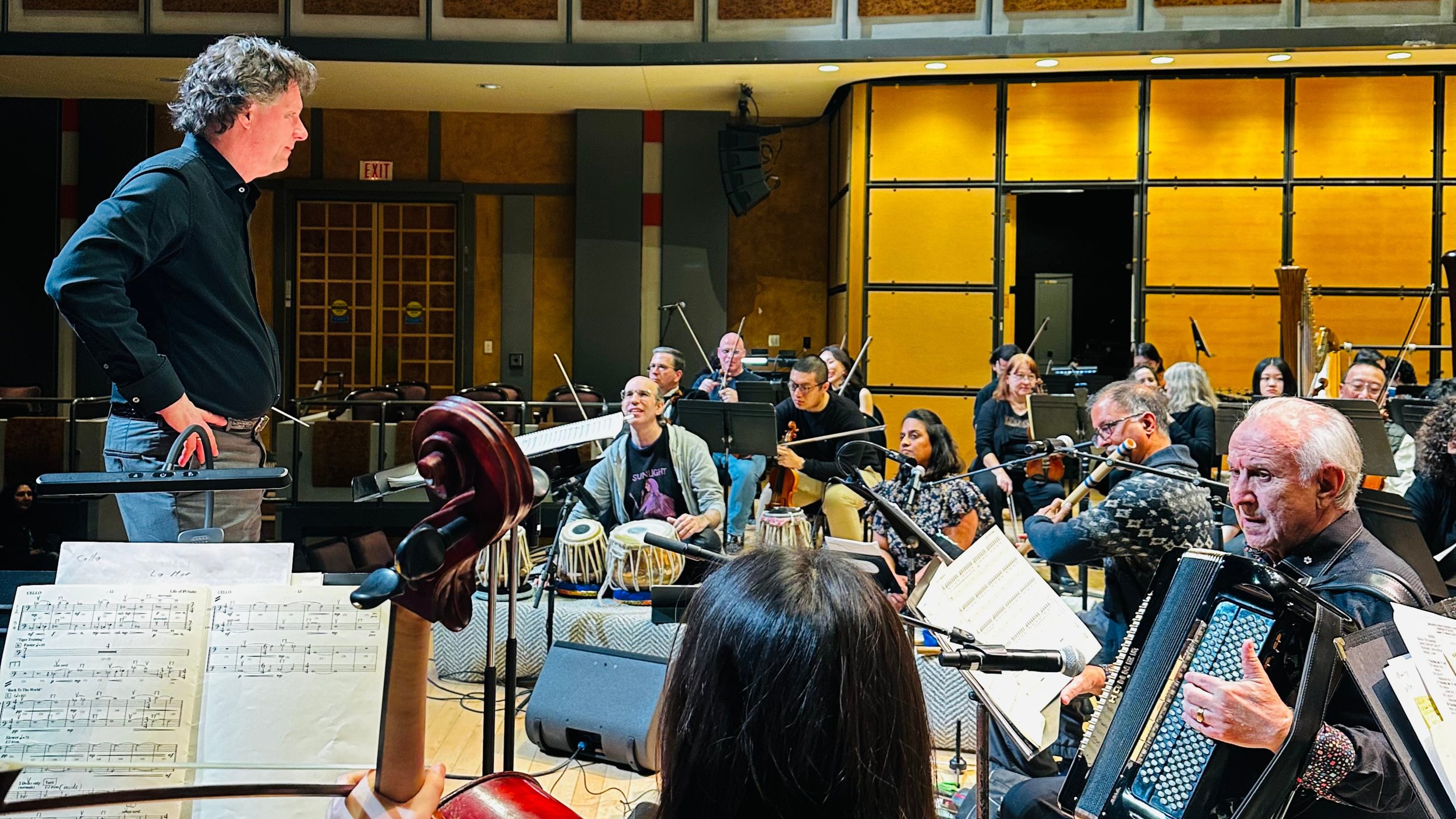A group of musicians in rehearsal, with a man standing at a music stand and others playing instruments such as a flute, accordion, and drums in a music studio or rehearsal space.