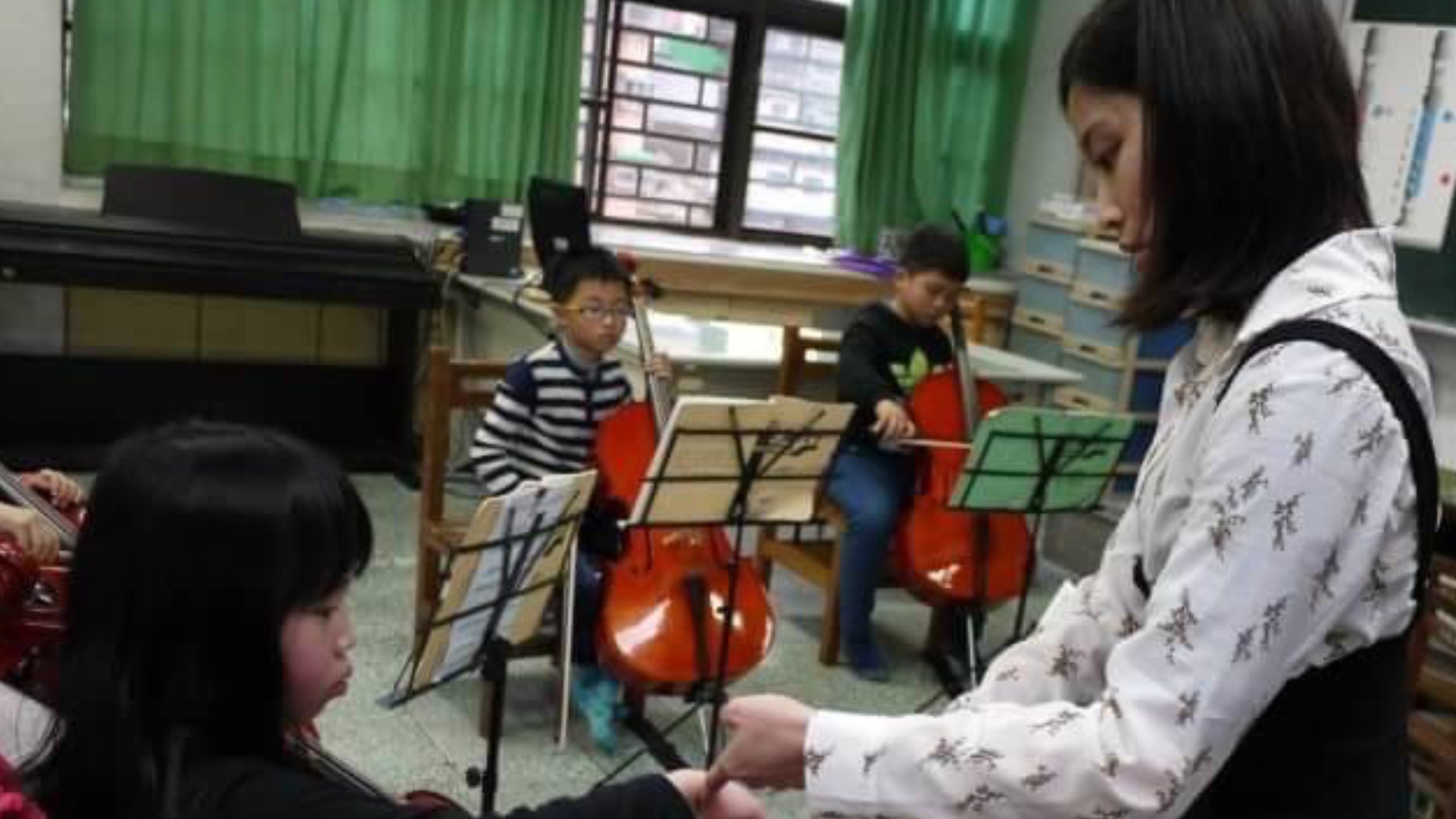 A music class with children playing cellos, a woman assisting, green curtains, and music stands in a classroom.