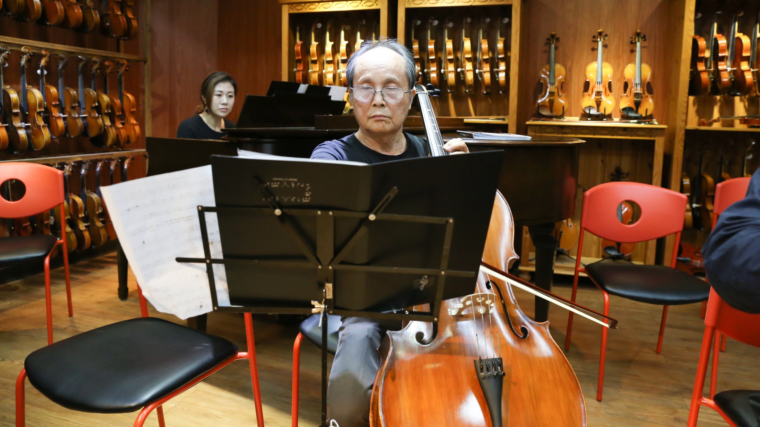 An elderly man with glasses playing the cello during a music rehearsal in a room with many violins on the wooden wall behind him. A woman is visible in the background at a piano.