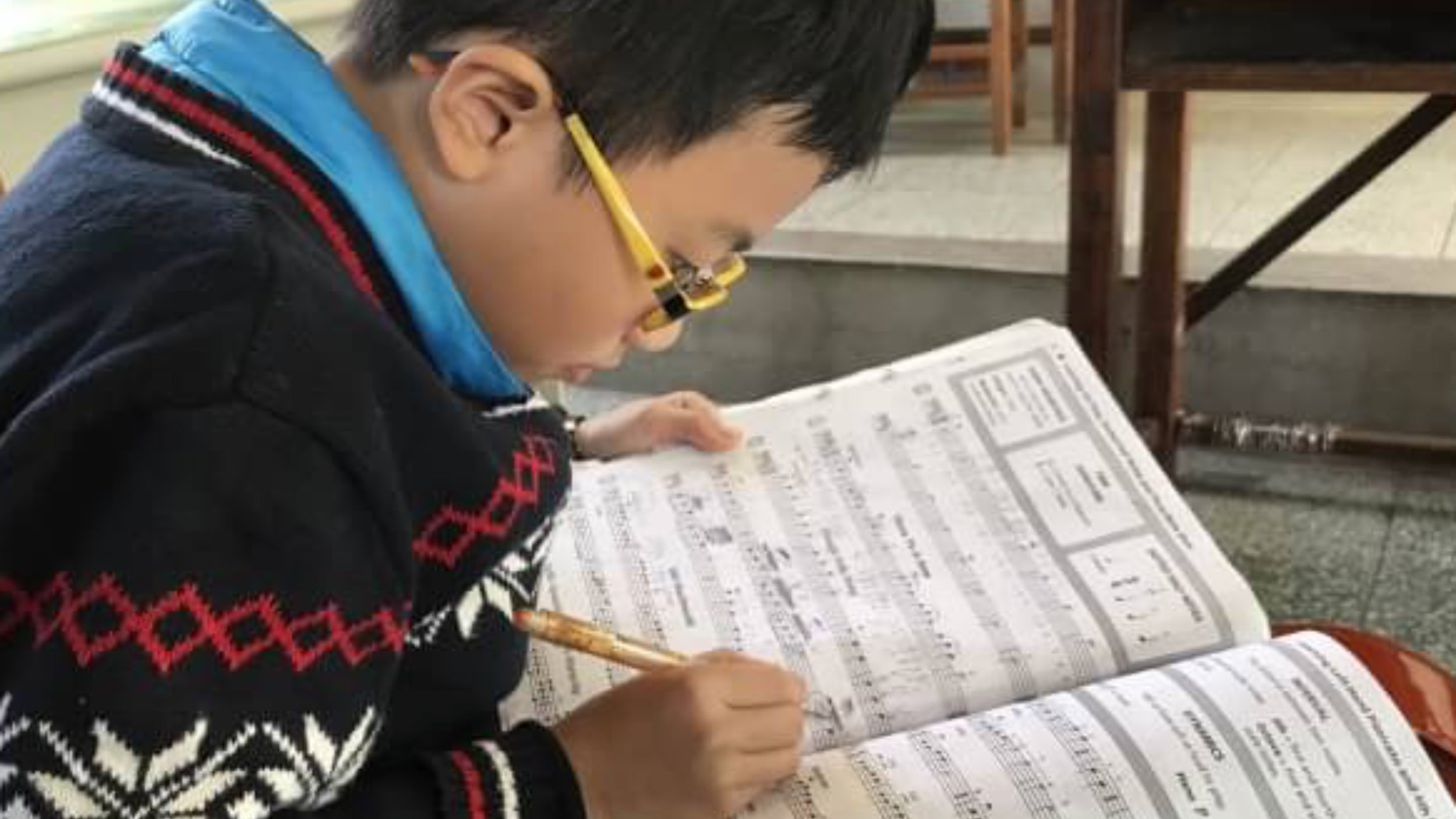 A young boy wearing glasses and a festive sweater sits at a table, reading sheet music and holding a pencil, with additional books or sheet music in front of him.