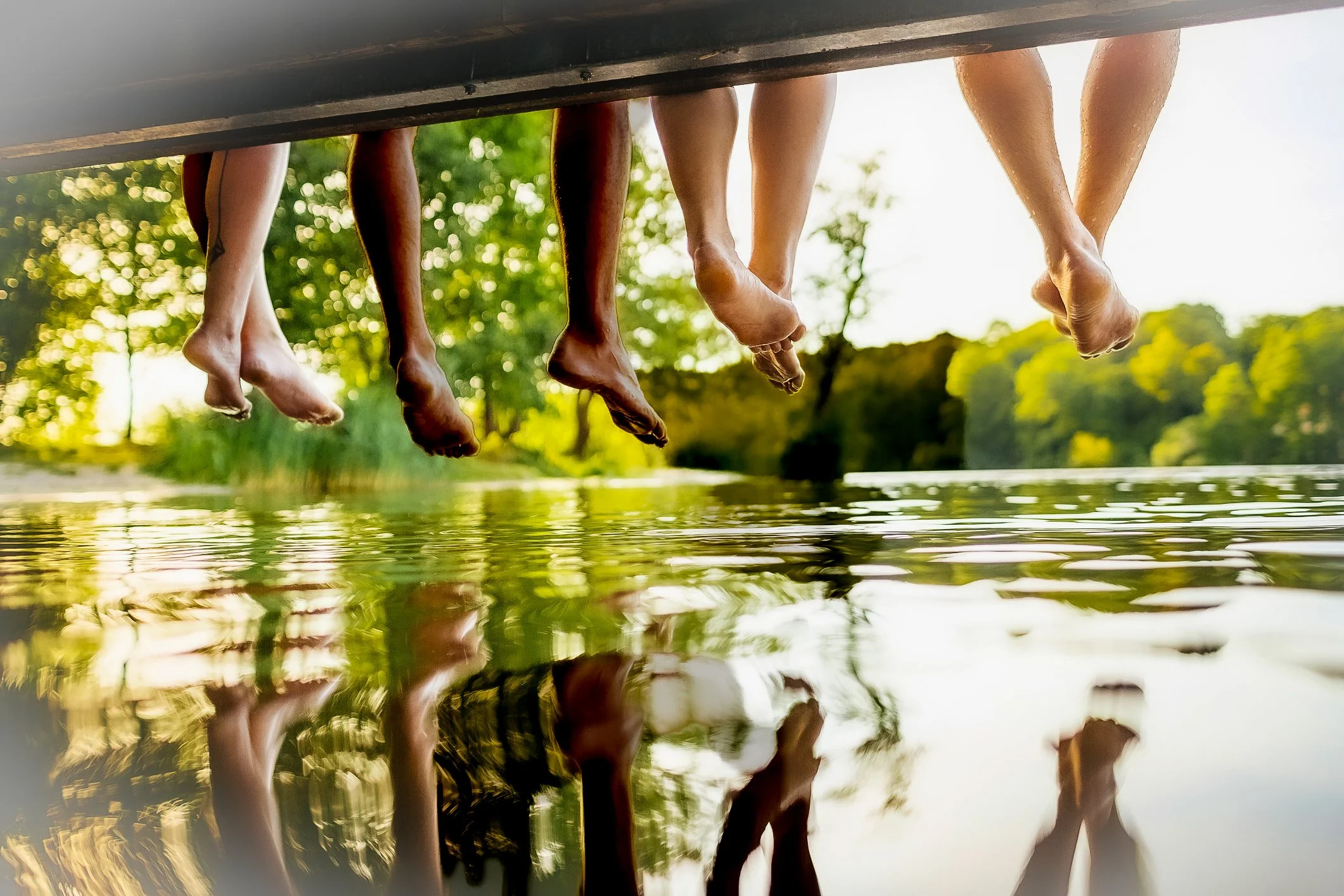 People sitting with their legs dangling over the edge of a dock, reflected in the water below, with trees and sunlight in the background.