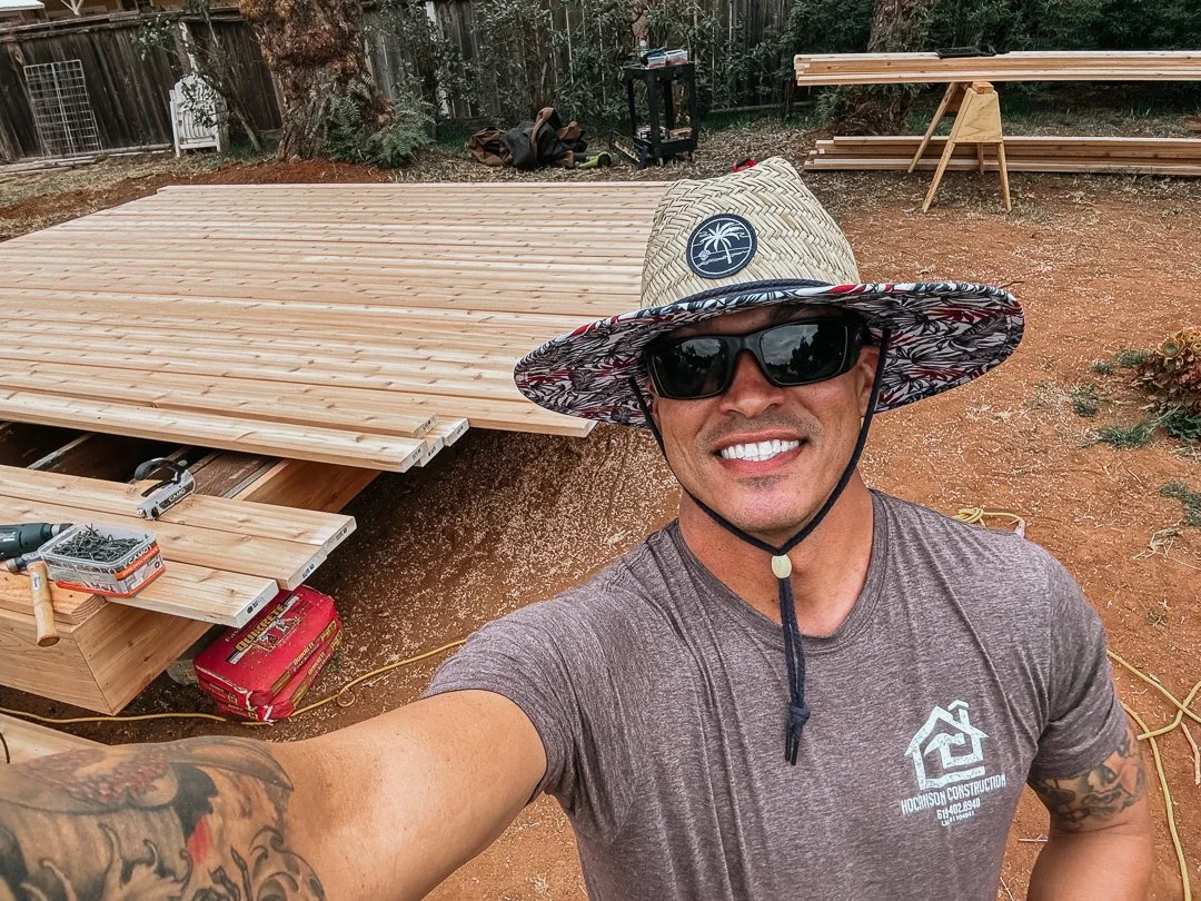 A man taking a selfie at a construction site with wooden planks, tools, and a partially built deck in the background.