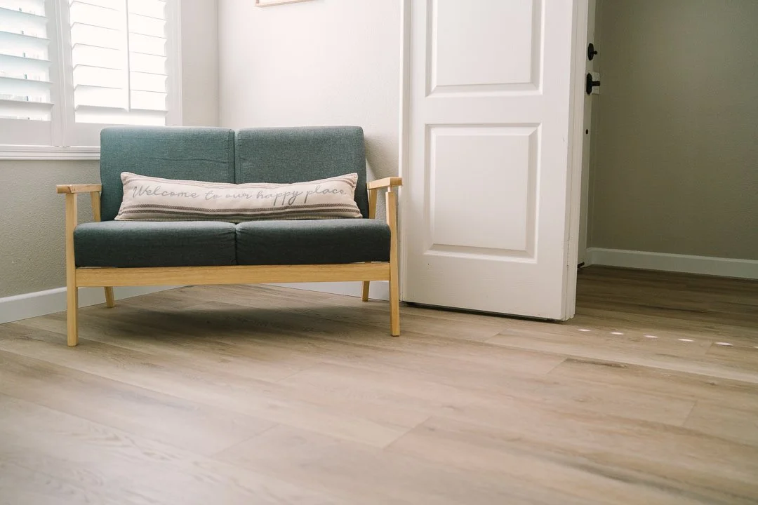 New floors look great in, A small teal sofa with wooden armrests and legs, a decorative pillow with the words 'Welcome to our happy place,'
