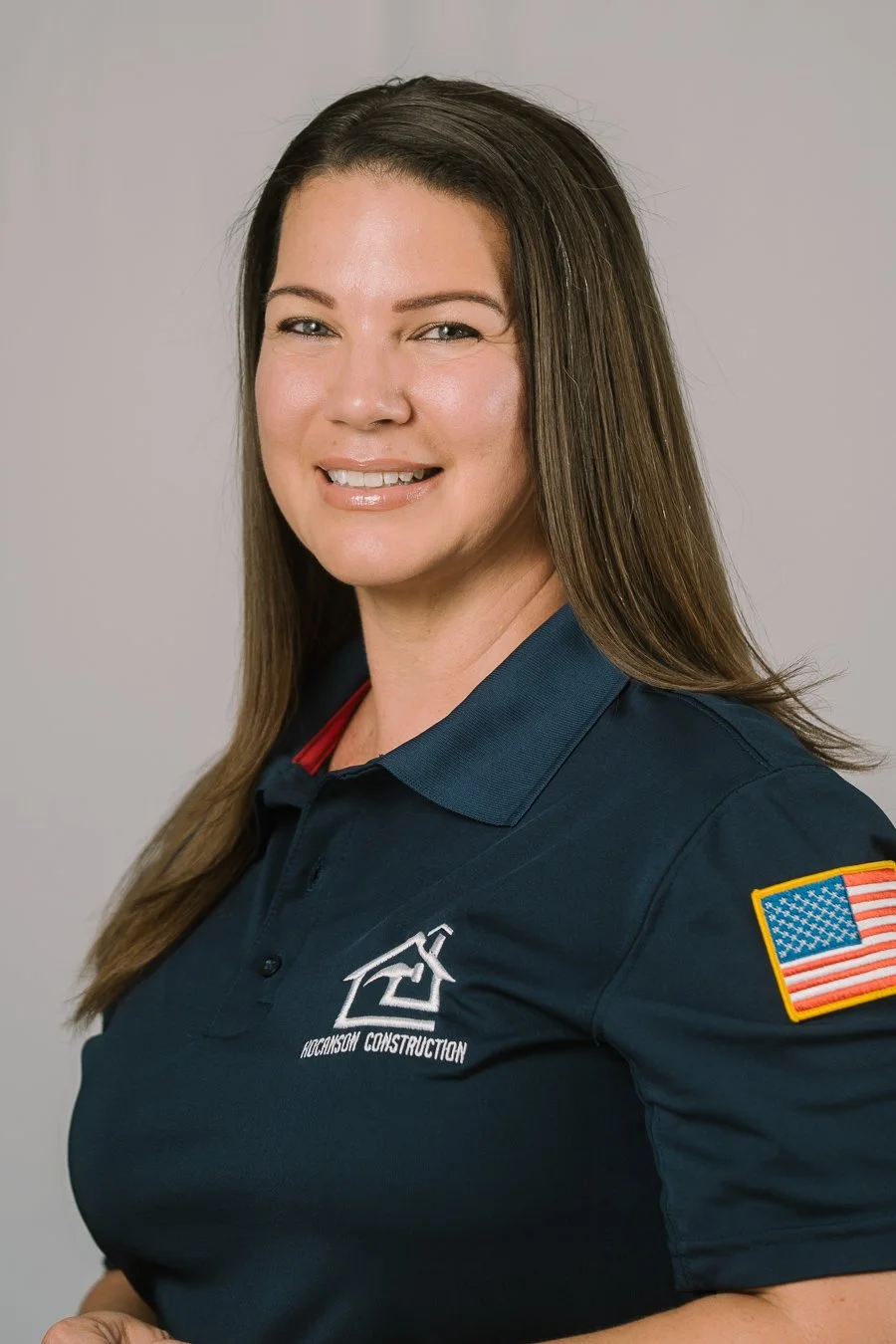 A woman with long brown hair, wearing a navy blue polo shirt with a logo reading 'Hockinson Construction' and an American flag patch on the sleeve. She is smiling and looking at the camera against a plain light gray background.