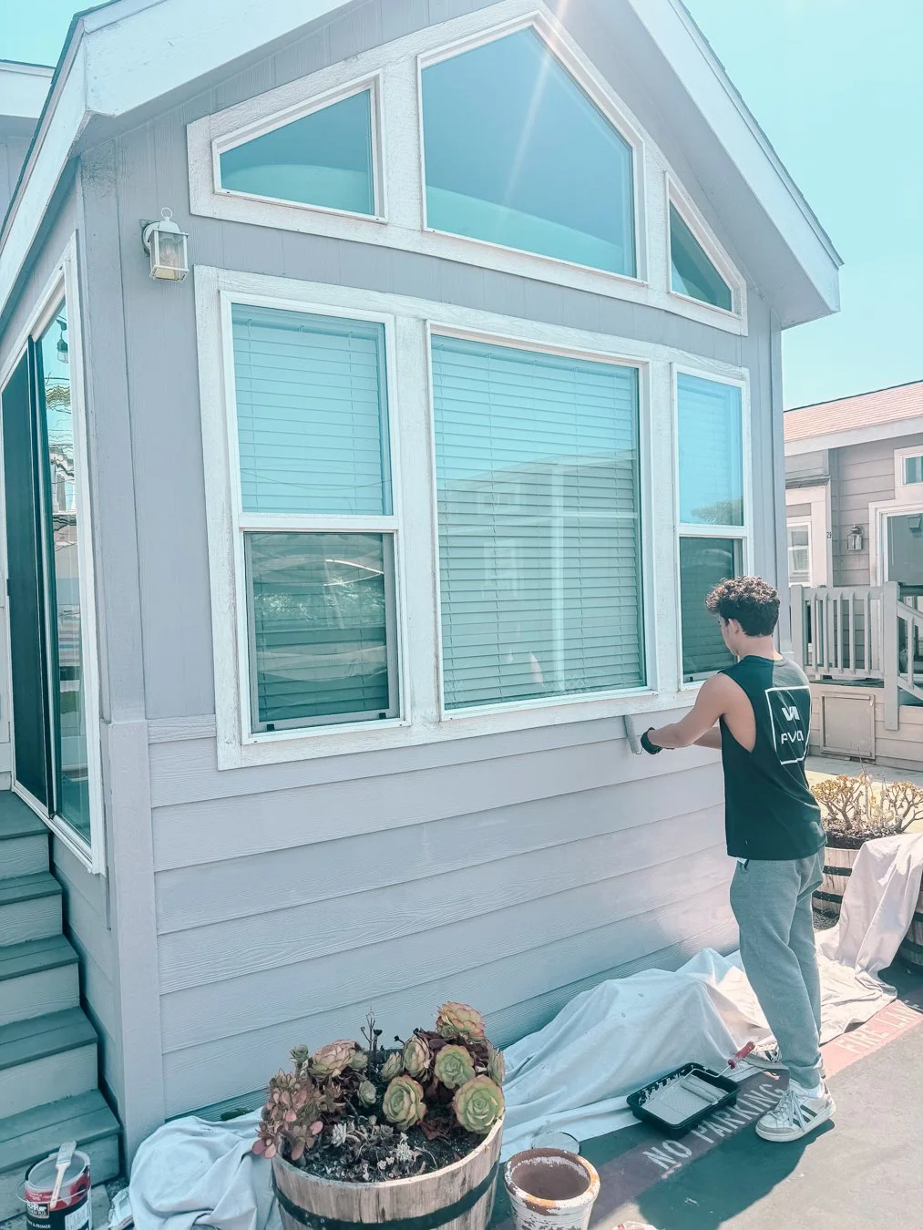 A person painting the exterior wall of a house with large windows on a sunny day.
