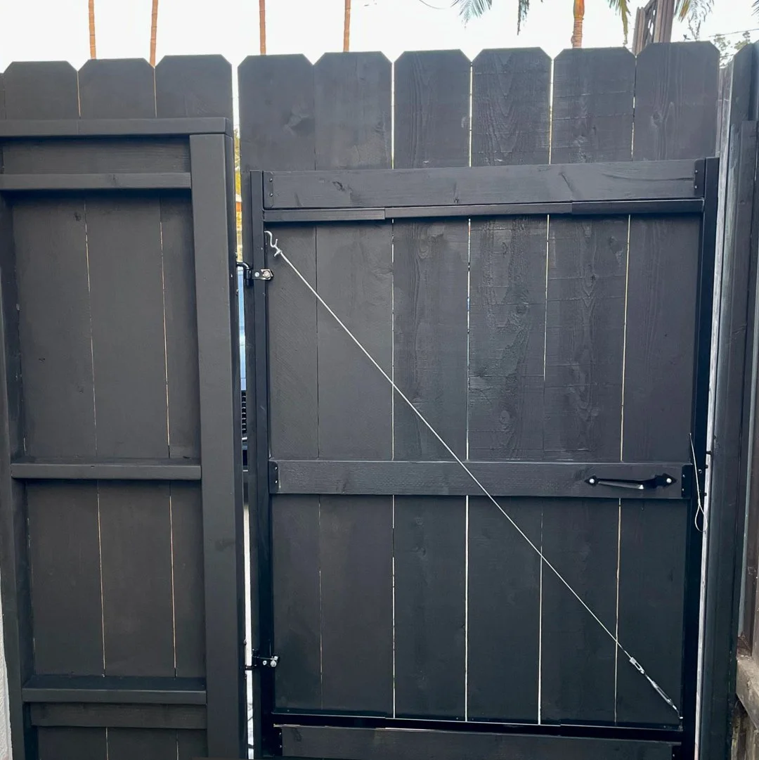Refinned fence, A black wooden gate with an angled top and a latch, attached to a matching black wooden fence, with a diagonal security cable.