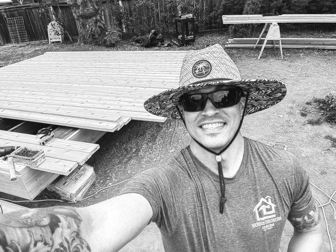 A man smiling, wearing sunglasses and a wide-brimmed straw hat with a palm tree logo, taking a selfie outdoors in a backyard with construction materials and a partially built wooden deck.