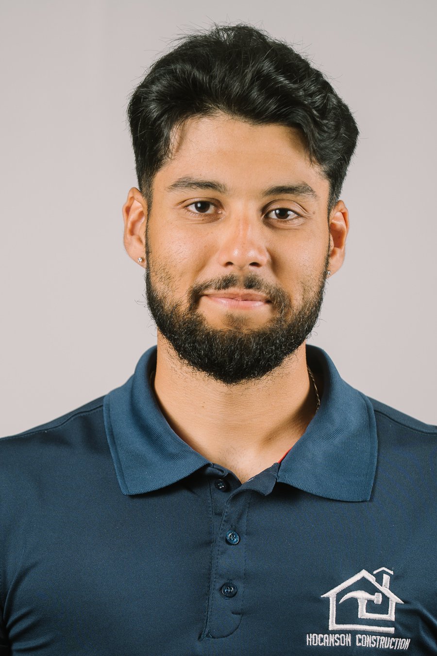 Portrait of a man with dark hair and beard, wearing a navy blue polo shirt with a construction company logo on it, against a plain gray background.
