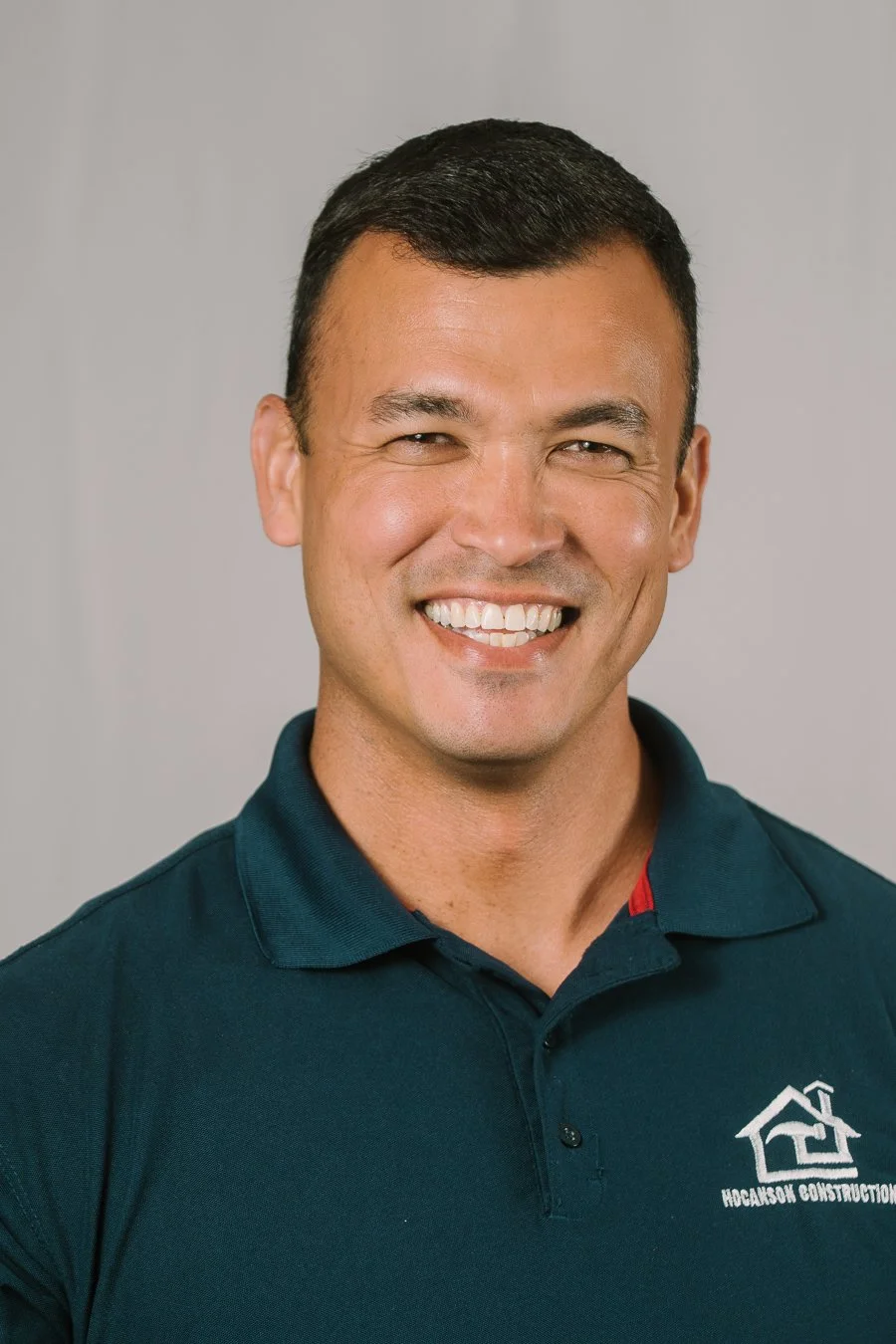 Smiling man in a dark blue polo shirt with a house logo from 'Hodgson Construction' on the chest, against a plain light gray background.