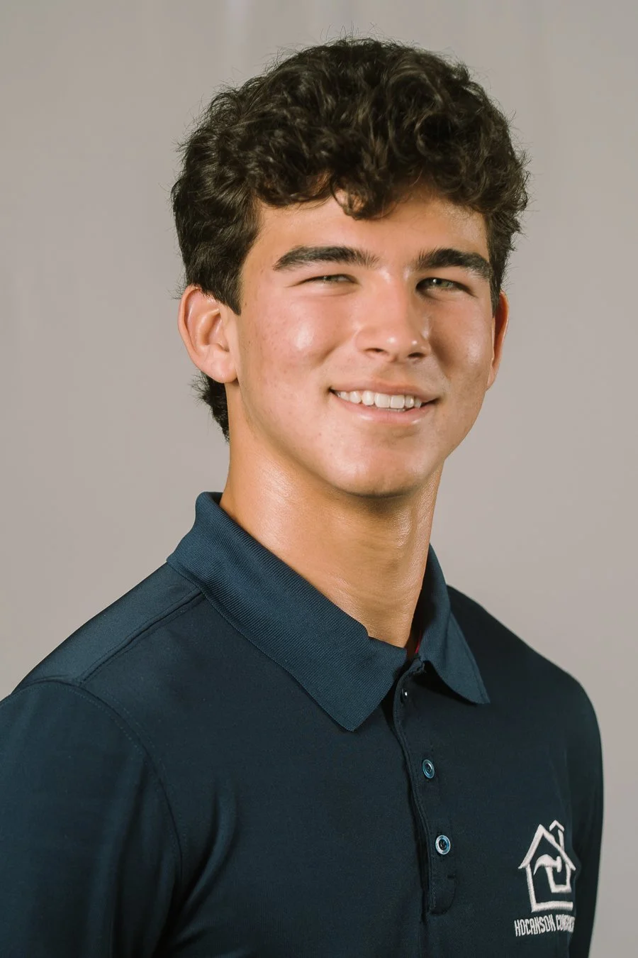 Young man with curly dark hair wearing a navy polo shirt with a logo on the chest, smiling at the camera against a plain gray background.