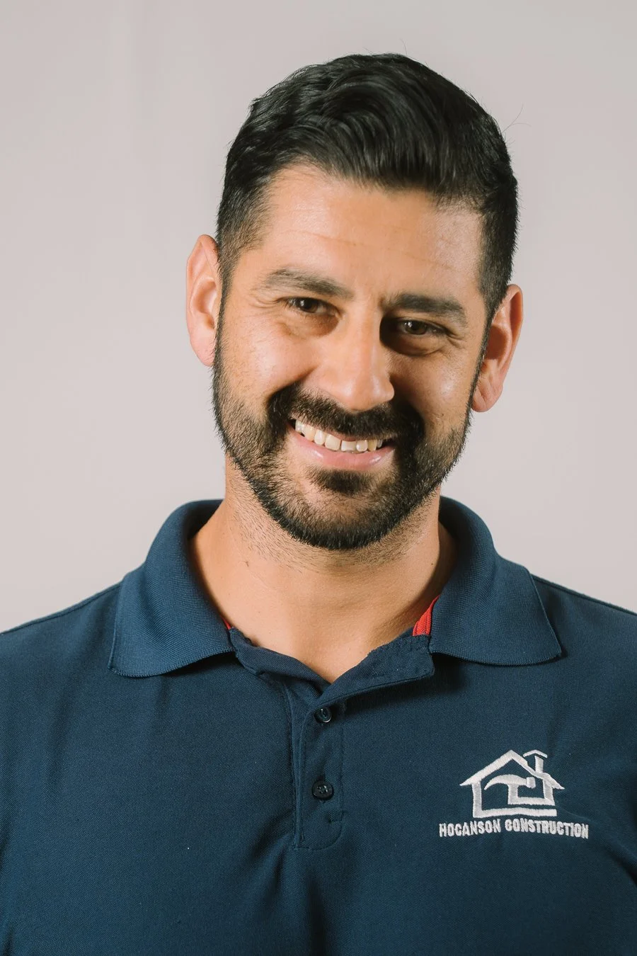 A smiling man with dark hair, beard, and mustache wearing a navy blue polo shirt with a logo that says "Hoganson Construction".