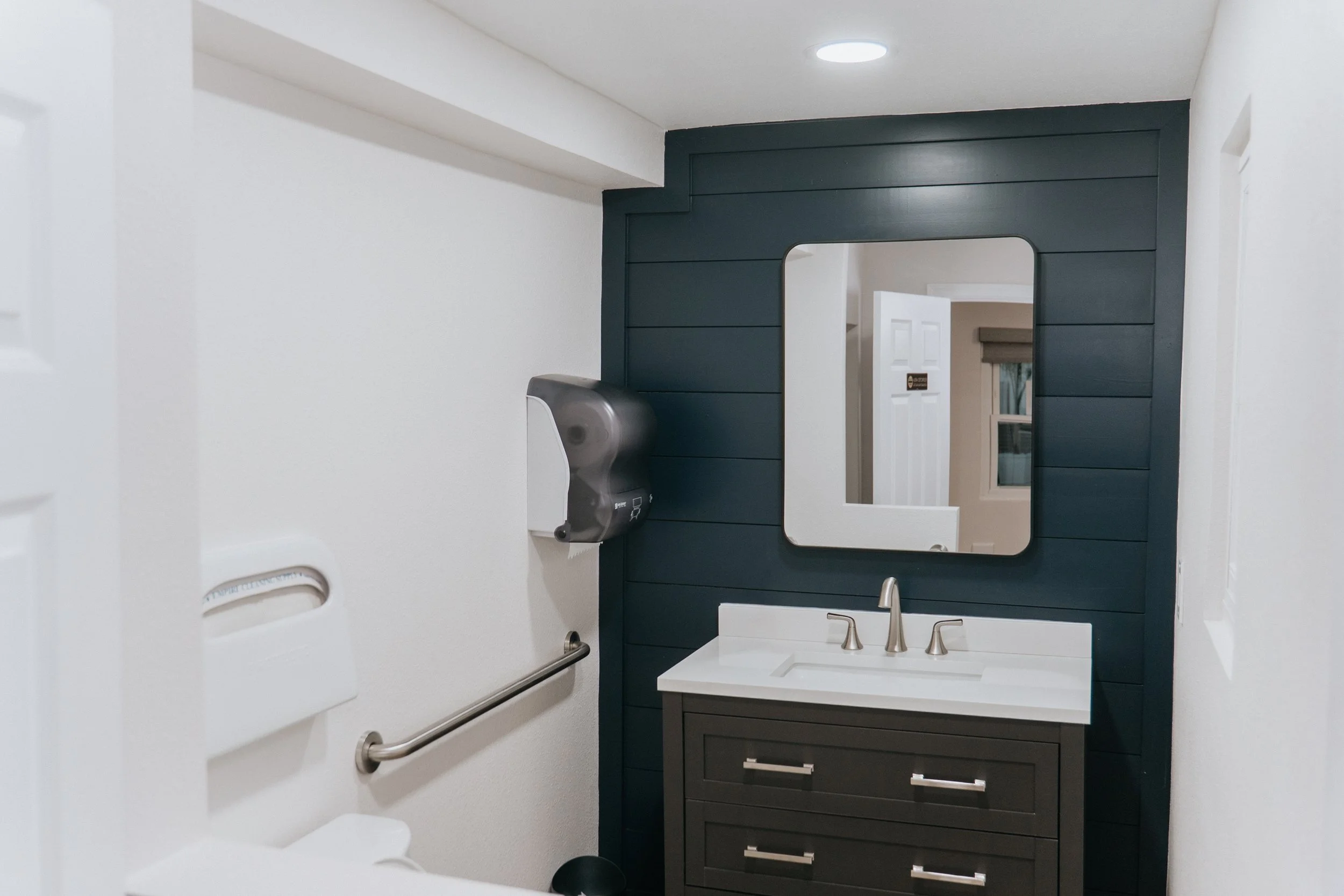 A fresh new bathroom fully remodeled. bathroom with a white sink, dark vanity cabinet, and a large mirror on a dark blue accent wall. The bathroom has a ceiling light, a hand dryer, and a window with a shade.