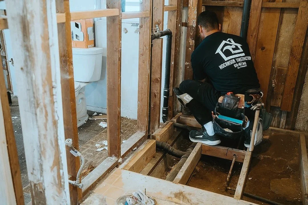 The boss, getting his hand A worker kneeling on a small platform installing plumbing in a bathroom under construction, with exposed wooden framing and a toilet in the background.
