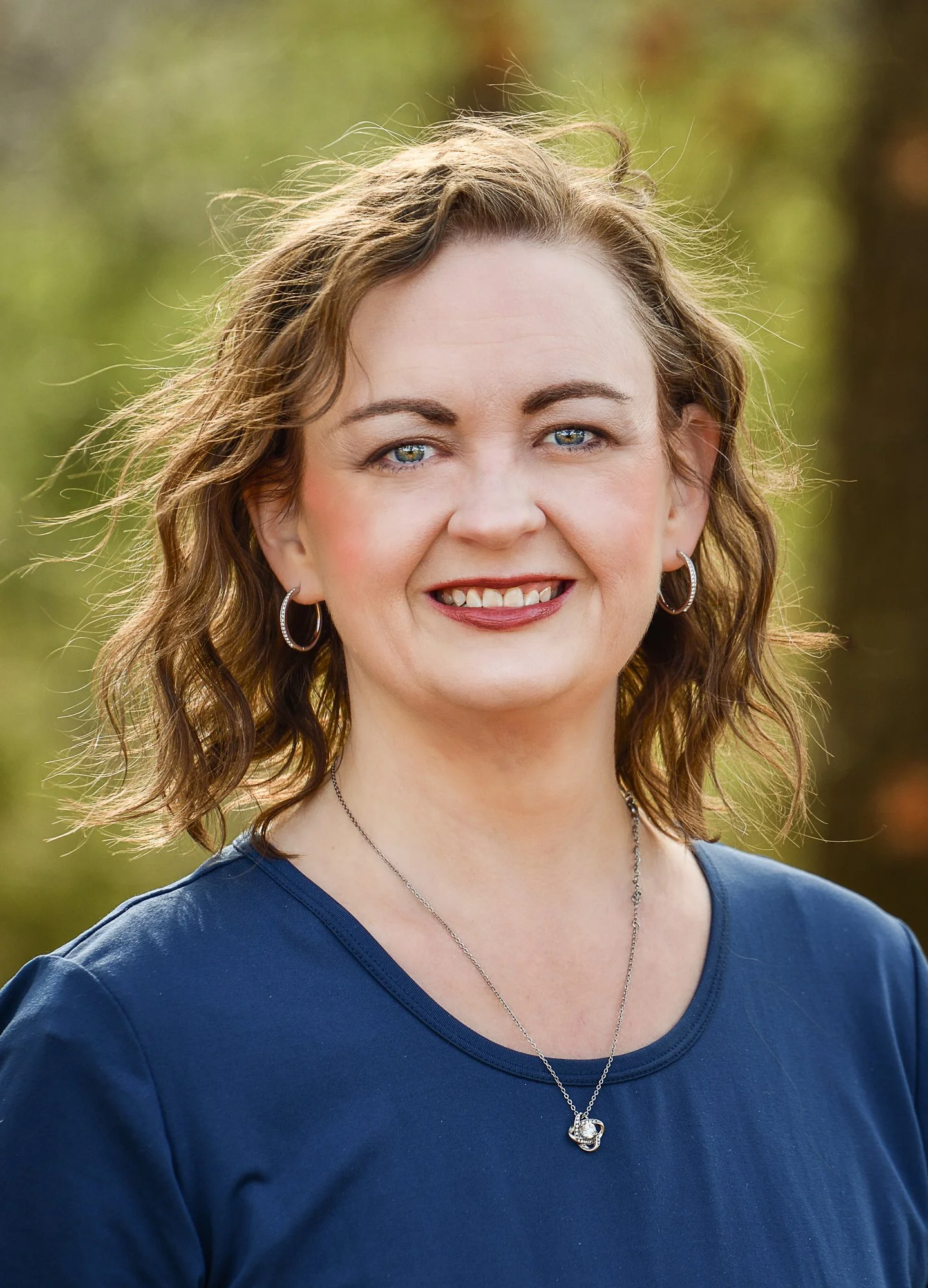 A woman smiling outdoors with trees in the background, wearing a blue shirt and silver jewelry.
