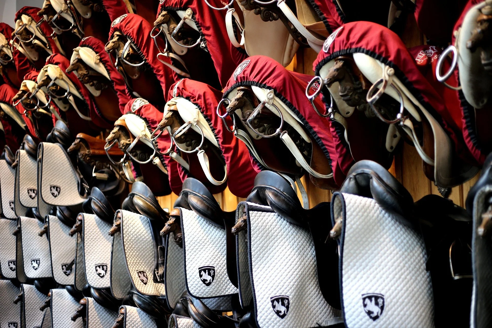 Rows of horse bridles, saddle pads, and horse helmets displayed in a tack shop, with the bridles and pads hanging on hooks and the helmets stacked below.