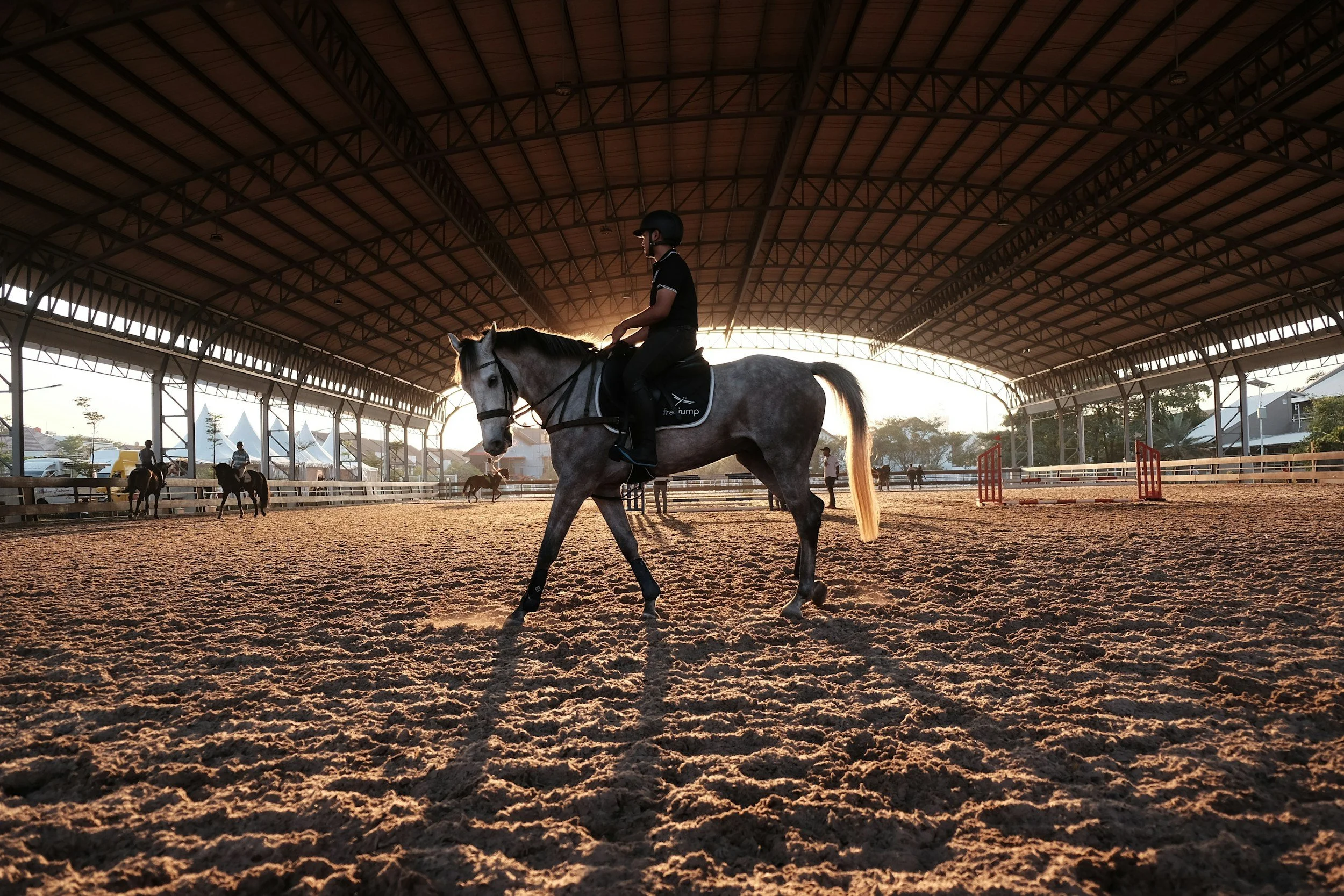 A person riding a gray horse inside an indoor riding arena during sunset, with other riders and horses visible in the background.