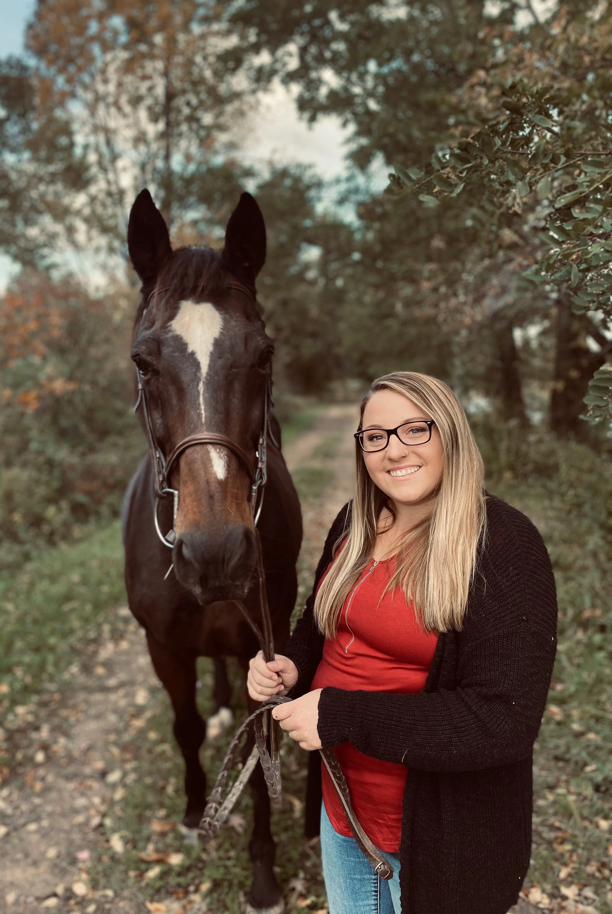 A woman with long blonde hair, glasses, and a red shirt, standing outdoors on a fall day, holding the lead of a dark brown horse with a white star on its forehead, against a background of autumn trees.