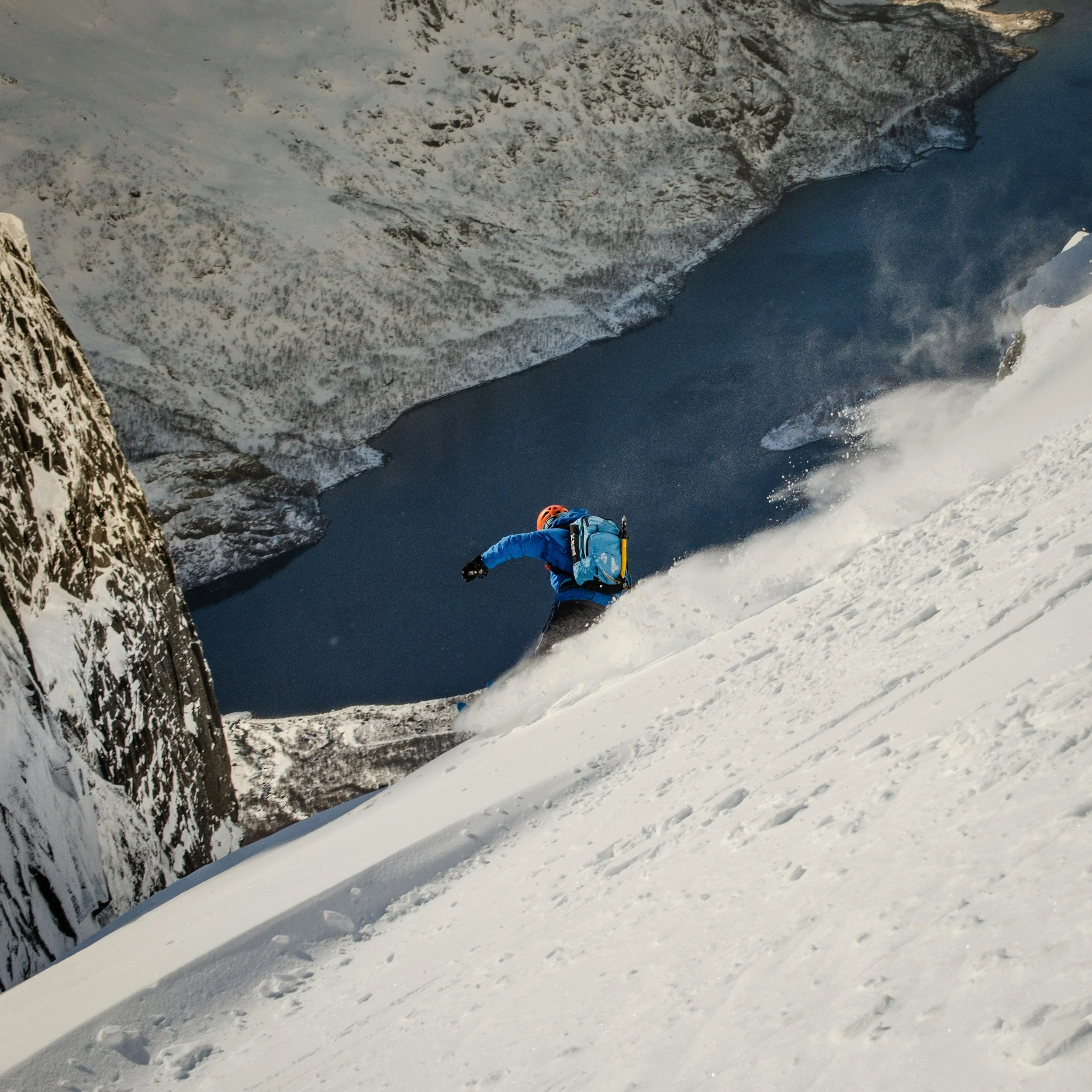 A person in blue winter gear and a helmet skiing down a snow-covered mountain slope, with a large body of water and snow-covered mountains in the background.