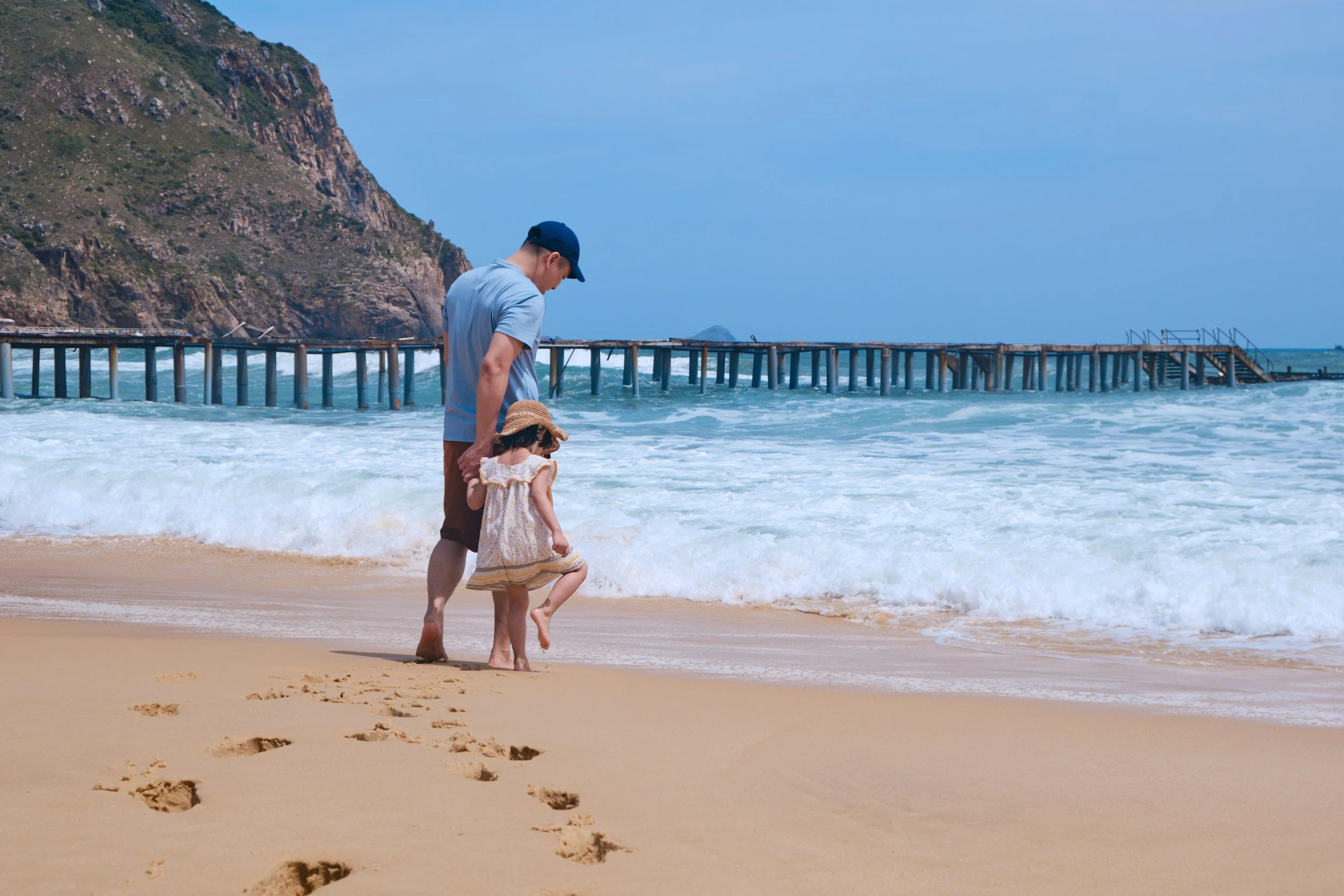 A man and a girl holding hands walking on the beach near the shoreline with footprints in the sand, a wooden pier extending into the ocean, and a rocky hillside under a blue sky.