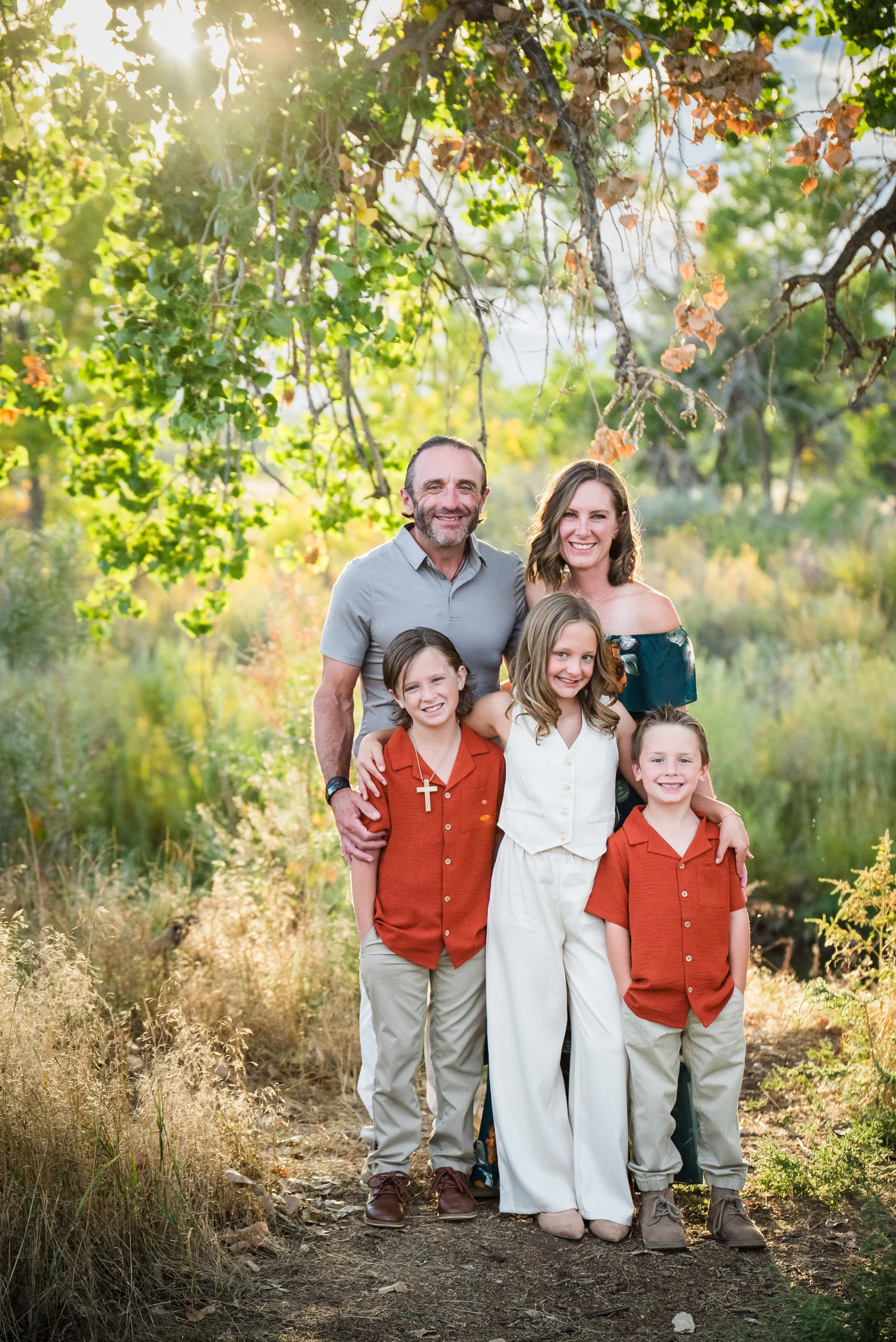 A family of six smiling and posing outdoors on a dirt trail surrounded by trees and greenery, with sunlight streaming through the leaves.