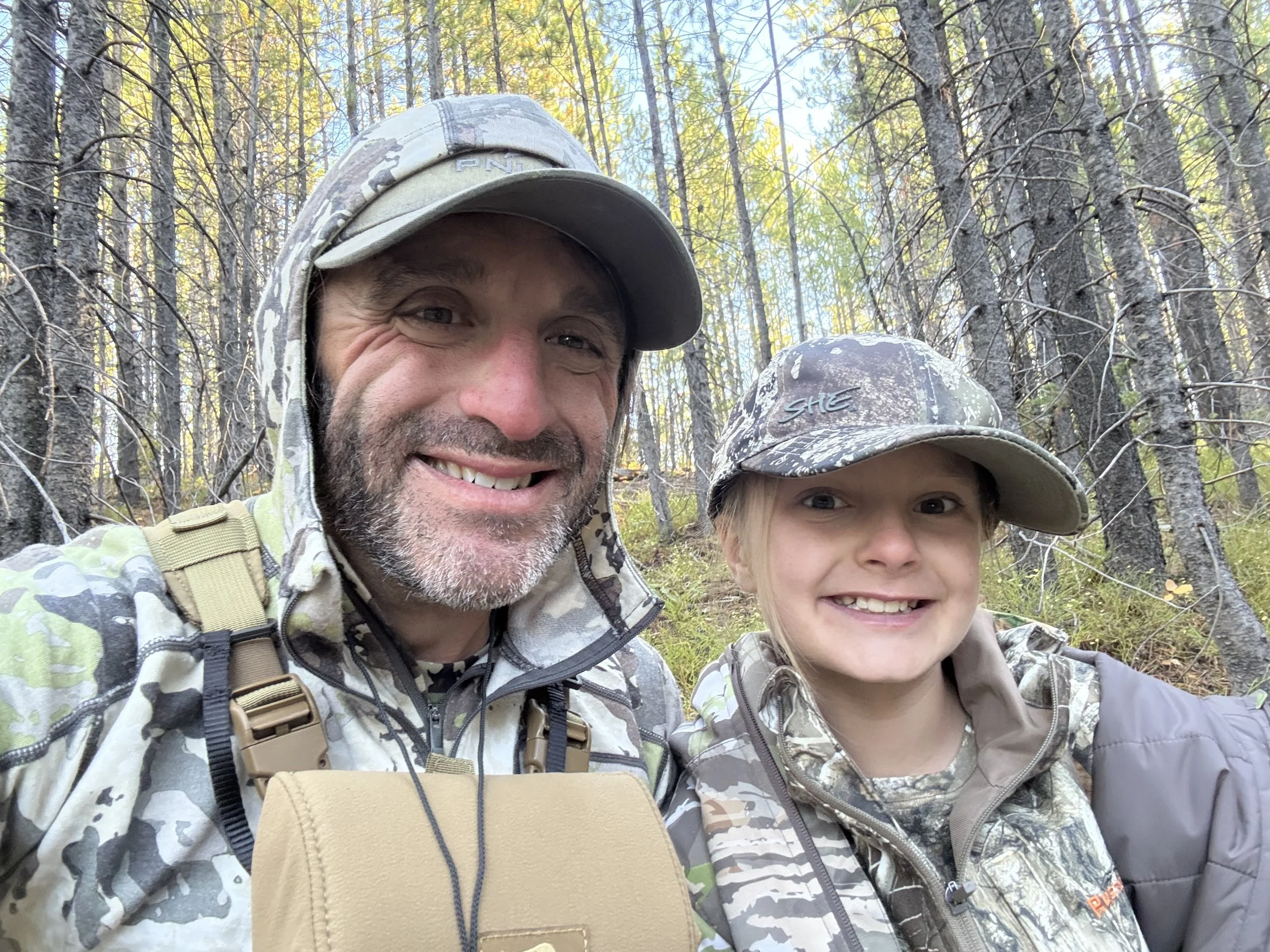 A man and a young girl smiling in a wooded area, both wearing camouflage clothing and hats, with trees and foliage in the background.