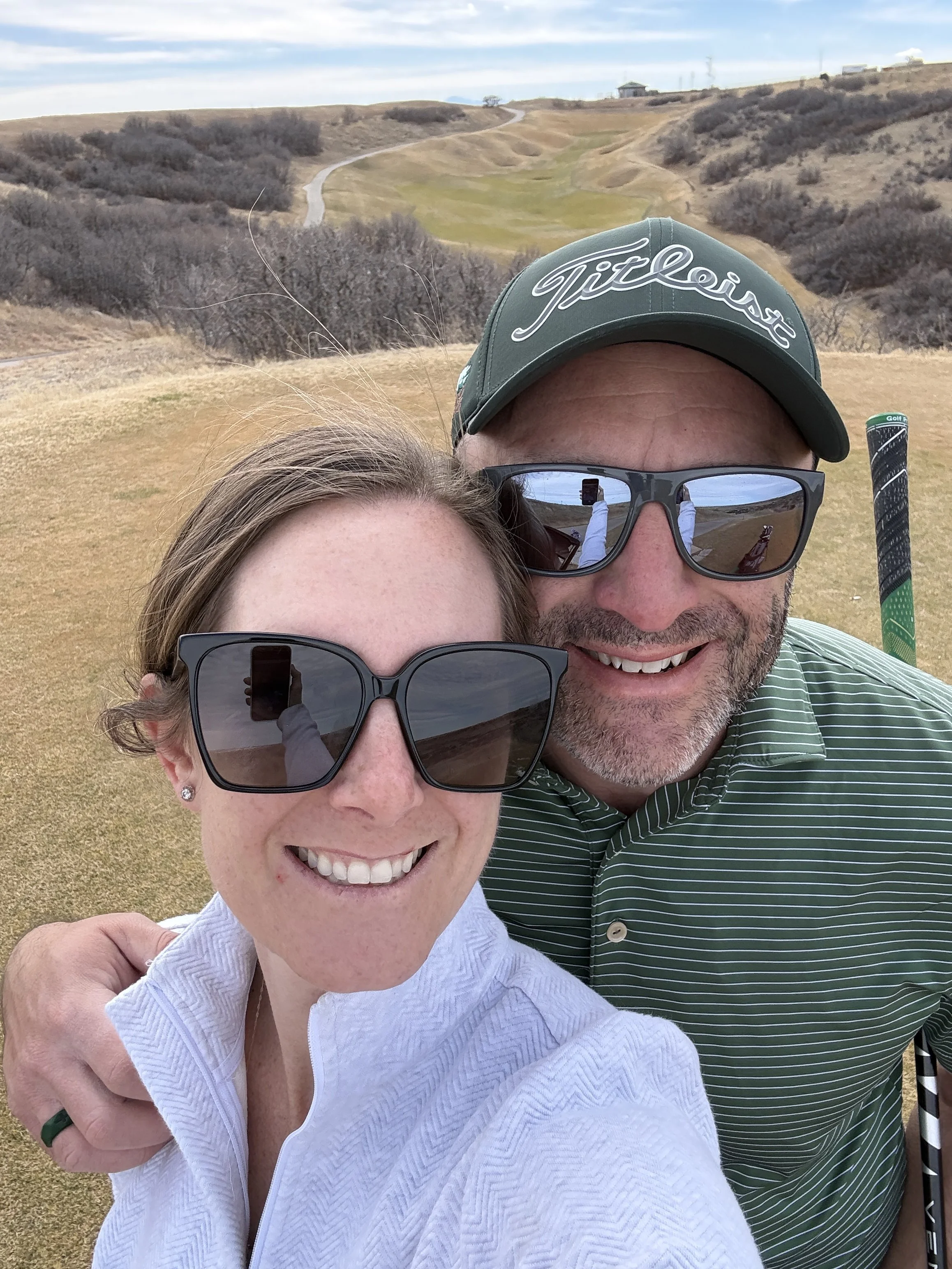 A smiling couple, a woman and a man, close together outdoors on a golf course. The woman is wearing sunglasses and a white top, and the man is wearing sunglasses, a green Titleist cap, and a striped polo shirt. They are taking a selfie with the golf course in the background.