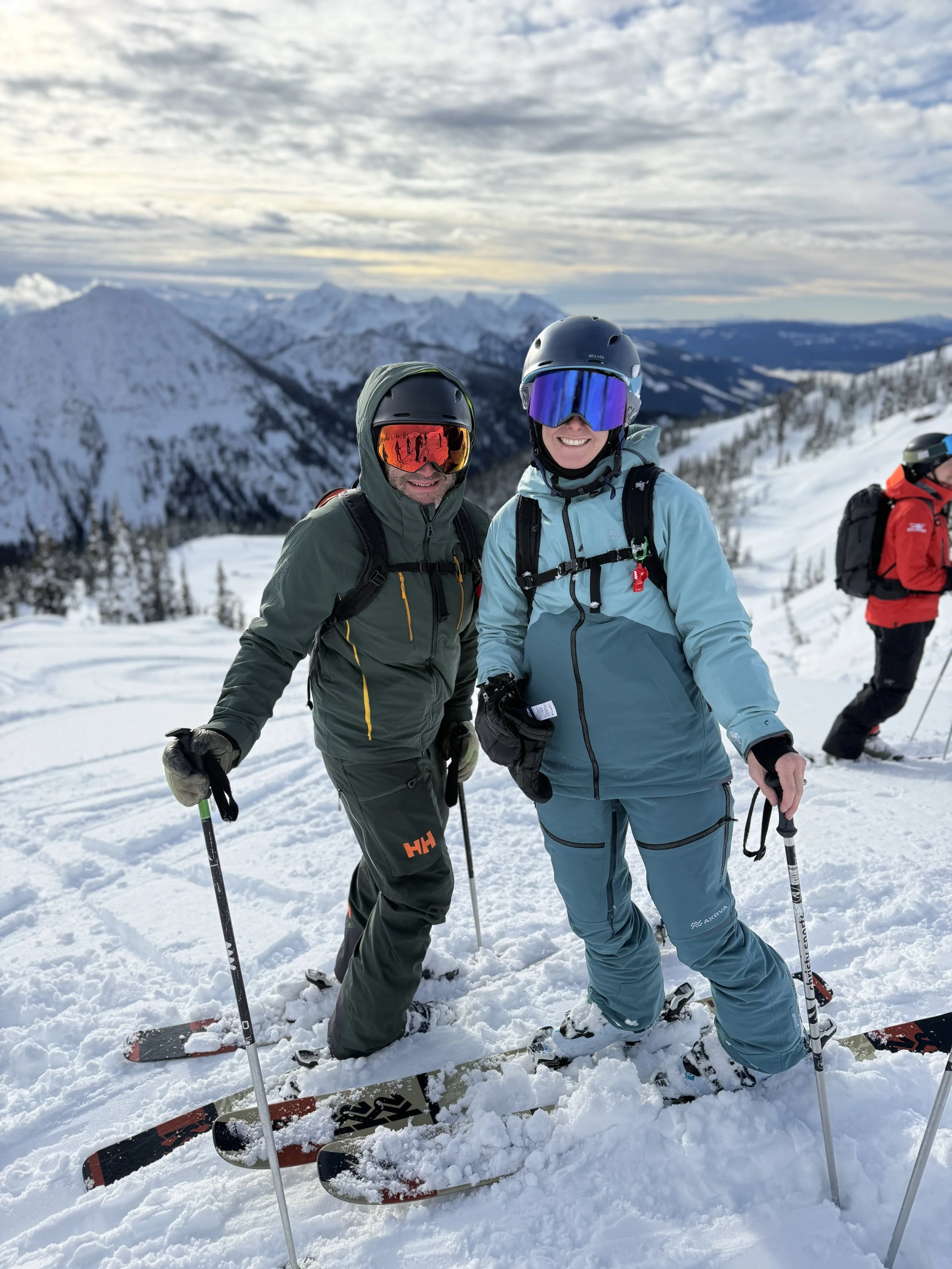 Two people in ski gear standing on snow-covered landscape with mountains and cloudy sky in the background. They are smiling and holding ski poles with skis at their feet. One person is wearing a green jacket and the other a blue jacket, both wearing helmets and goggles.