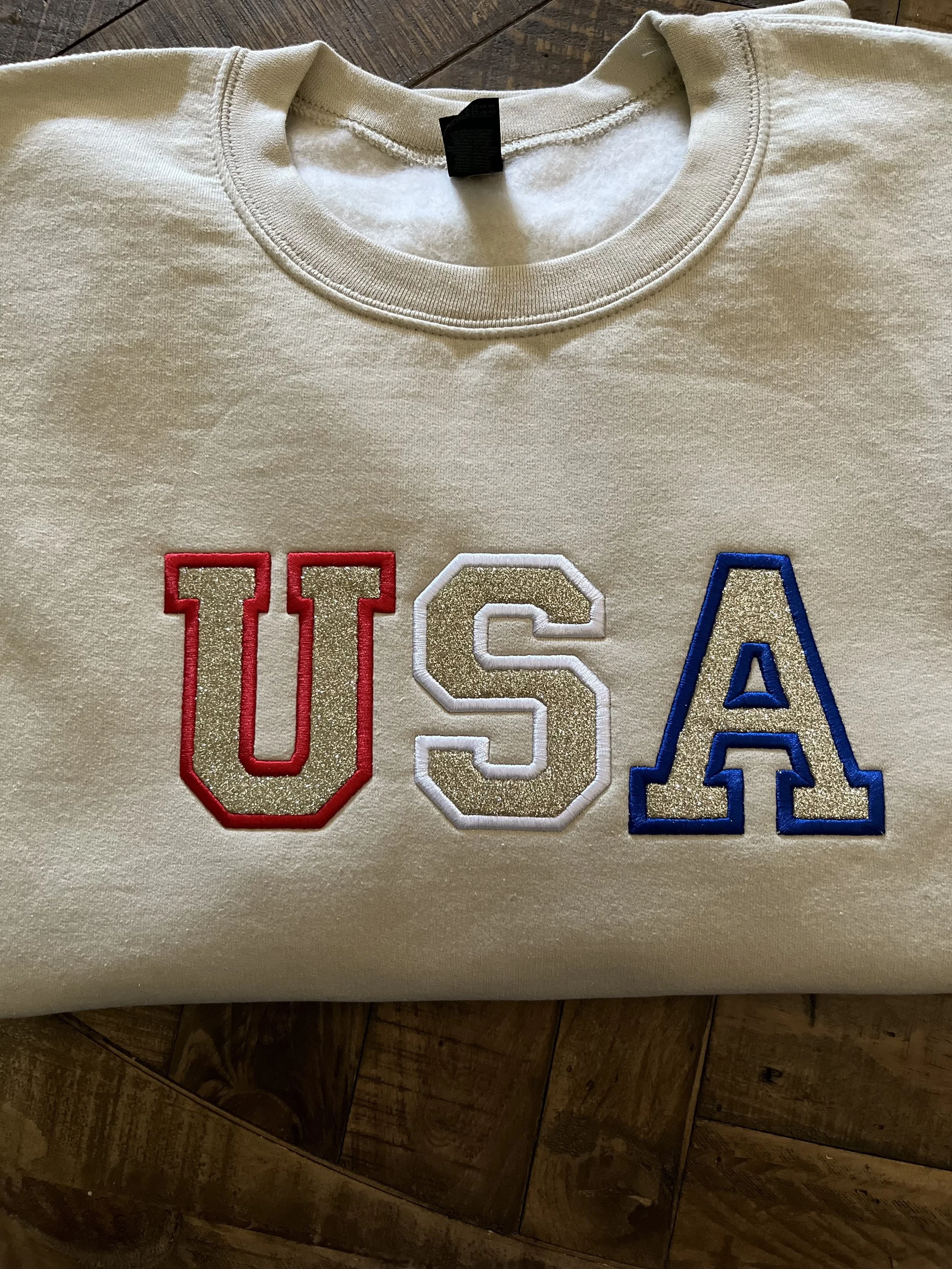 Cream-colored sweatshirt with embroidered letters U, S, A in red, white, and blue respectively, placed on a wooden surface.