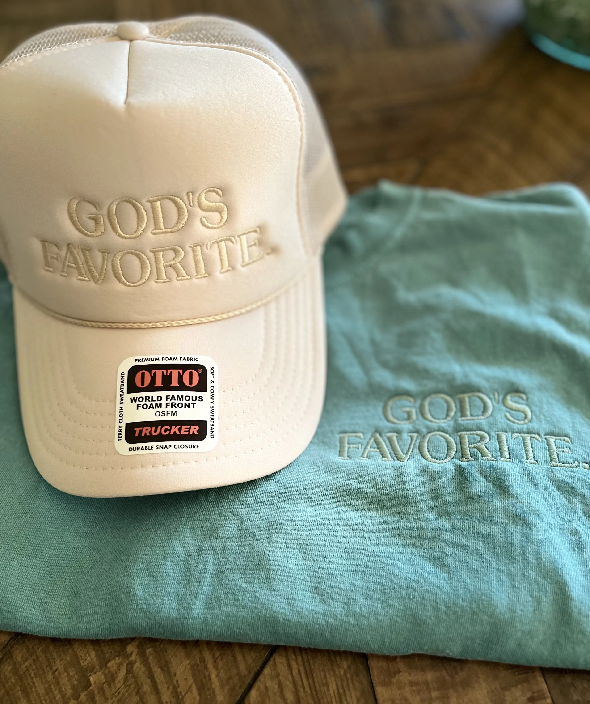 A beige baseball cap with embroidered text that reads 'GOD'S FAVORITE' and a part of a light blue cloth with similar embroidered text. The items are on a hardwood floor.