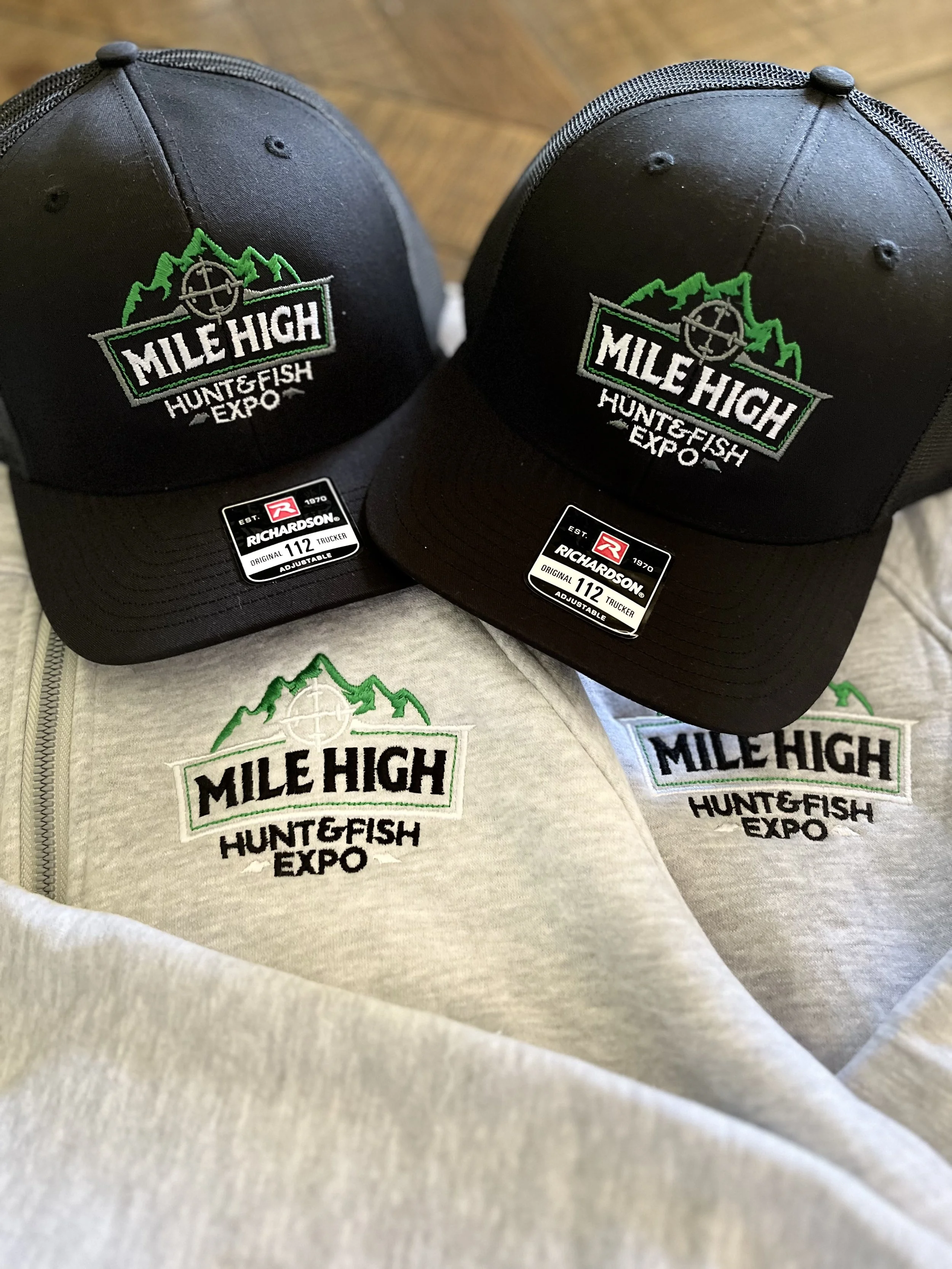 Two black caps with embroidered logo and text for the Mile High Hunt Fish Expo, placed on a hoodie and a fabric surface, with stickers indicating size and brand.