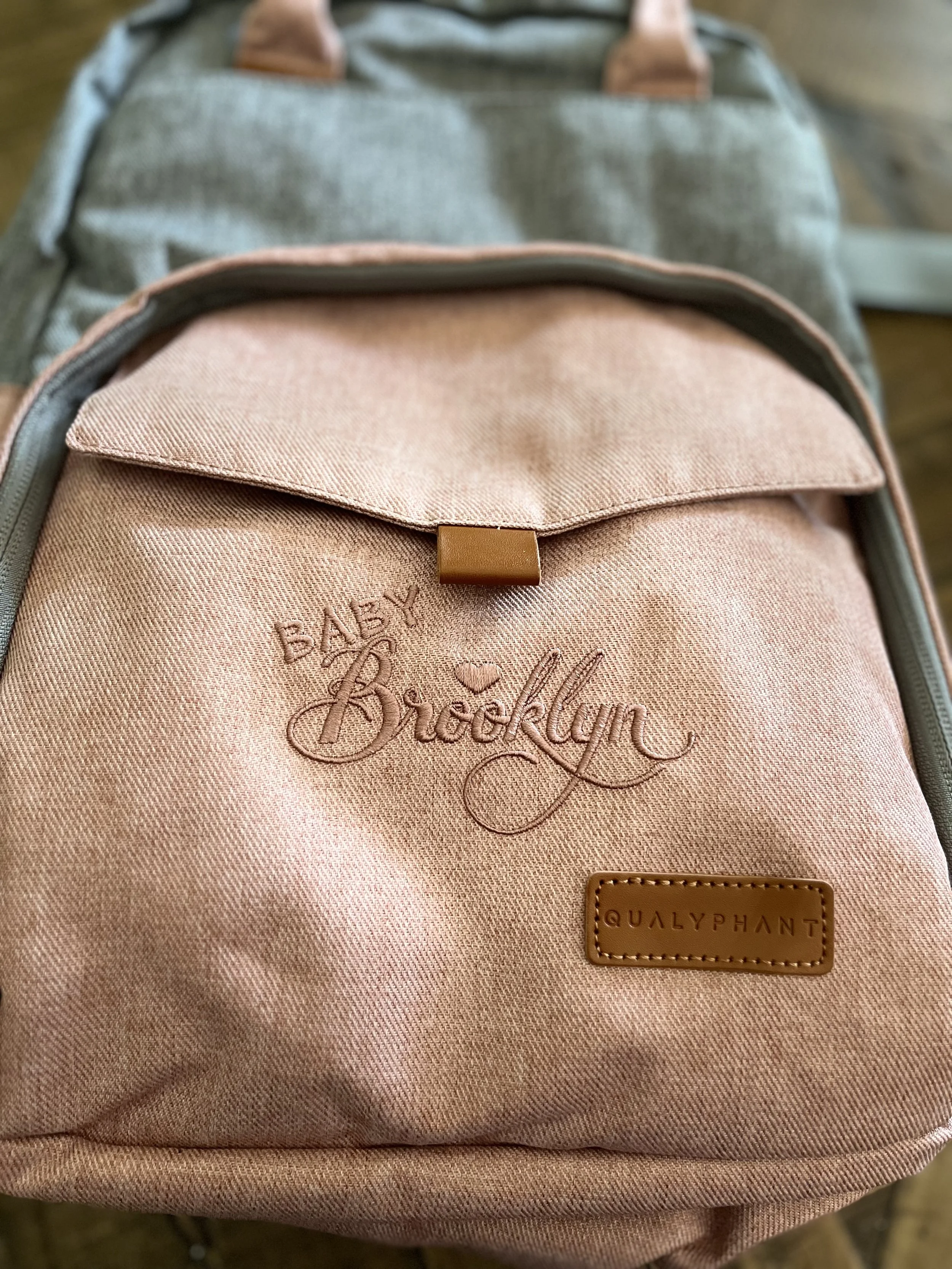 A pink backpack with embroidered text that reads 'Baby Brecklyn' and a small leather patch with the brand name '@QualPhant'.