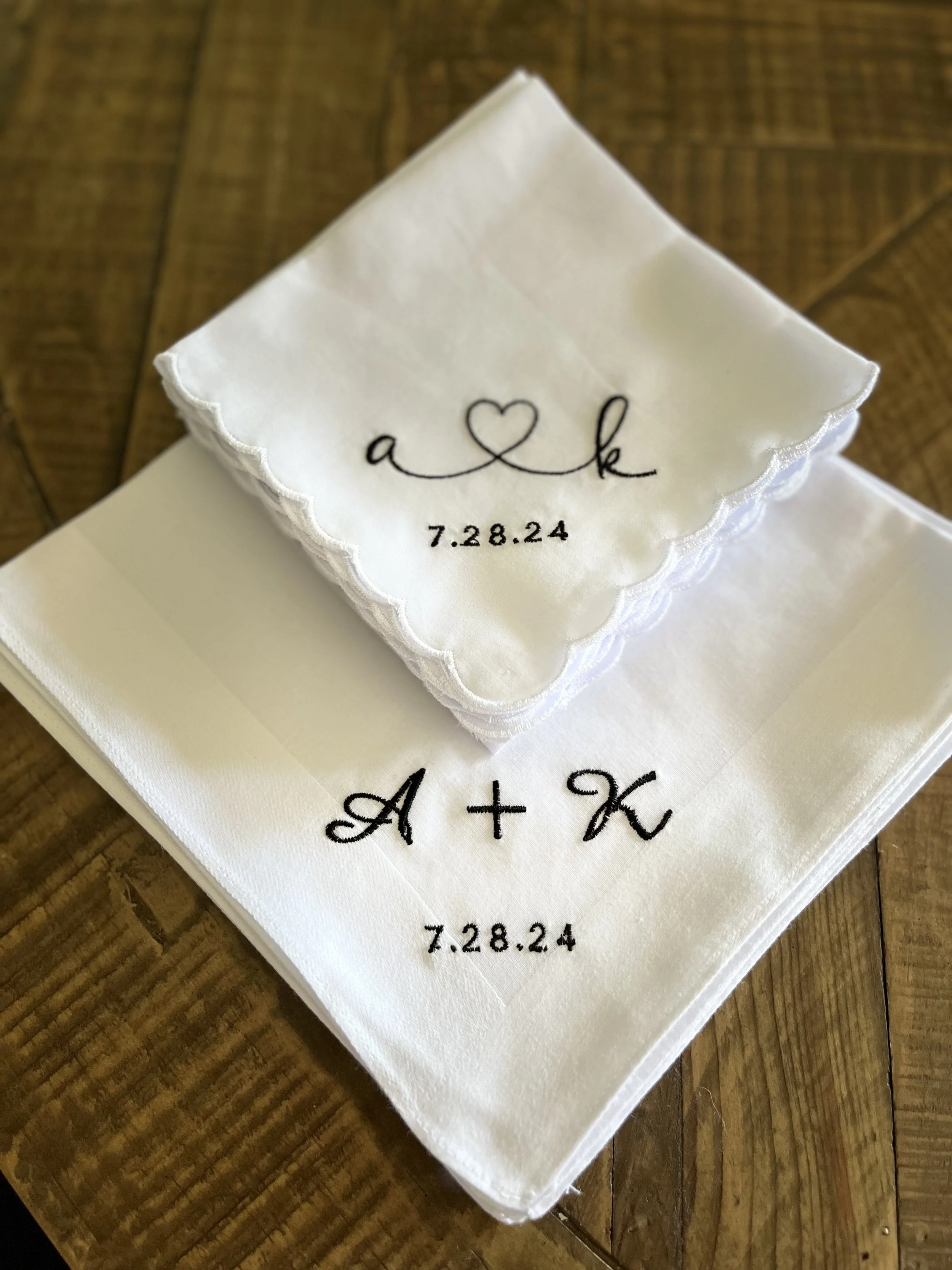 Close-up of a white satin ring bearer pillow with embroidery of the word 'a' a heart symbol, 'k' and the date '7.28.24'. The pillow is placed on a wooden surface.