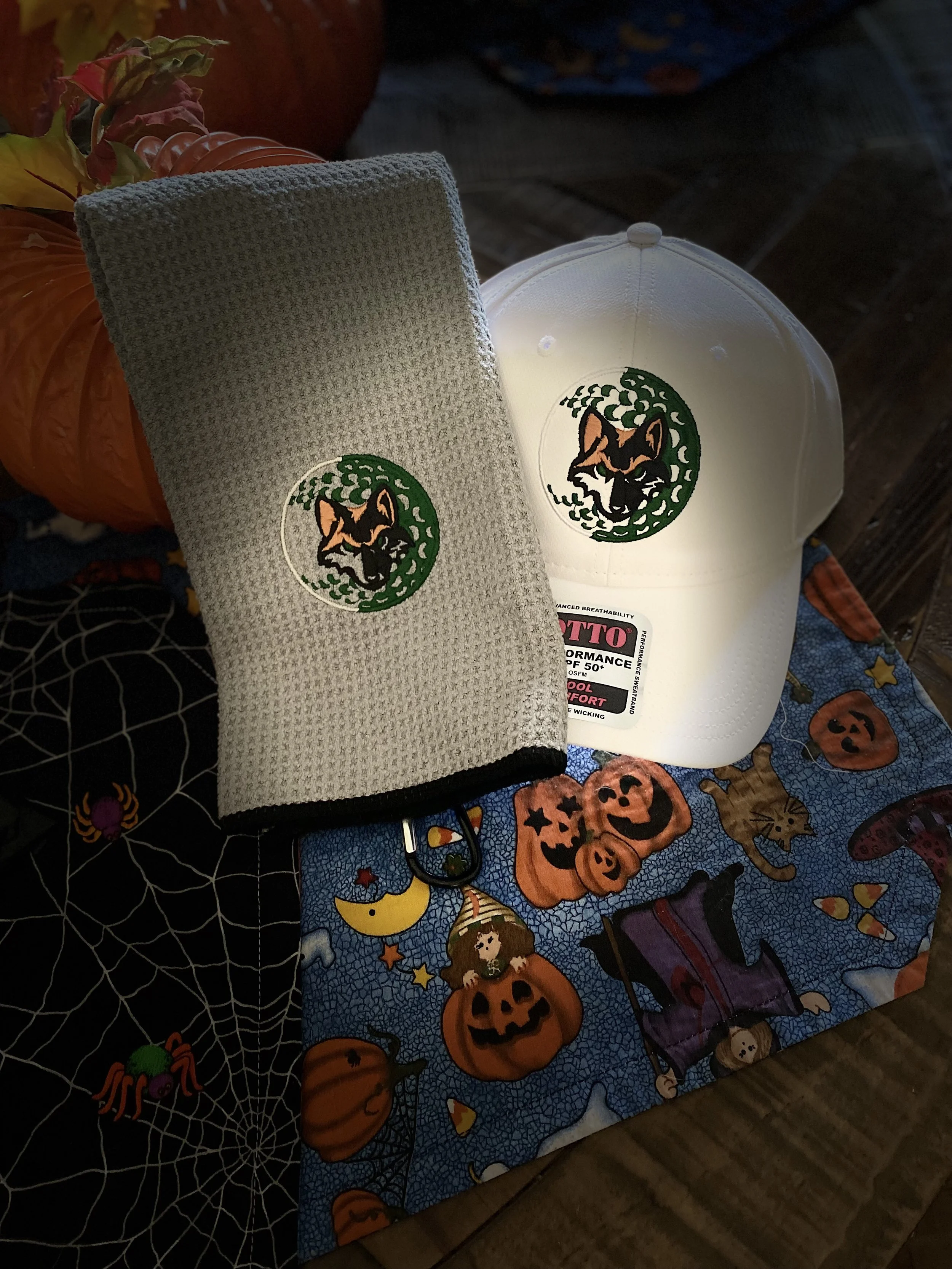 Halloween-themed tablecloth with pumpkin, ghost, and spider designs, a white cap with a logo, a gray headband with a logo, and a pumpkin decoration surrounded by fall leaves.