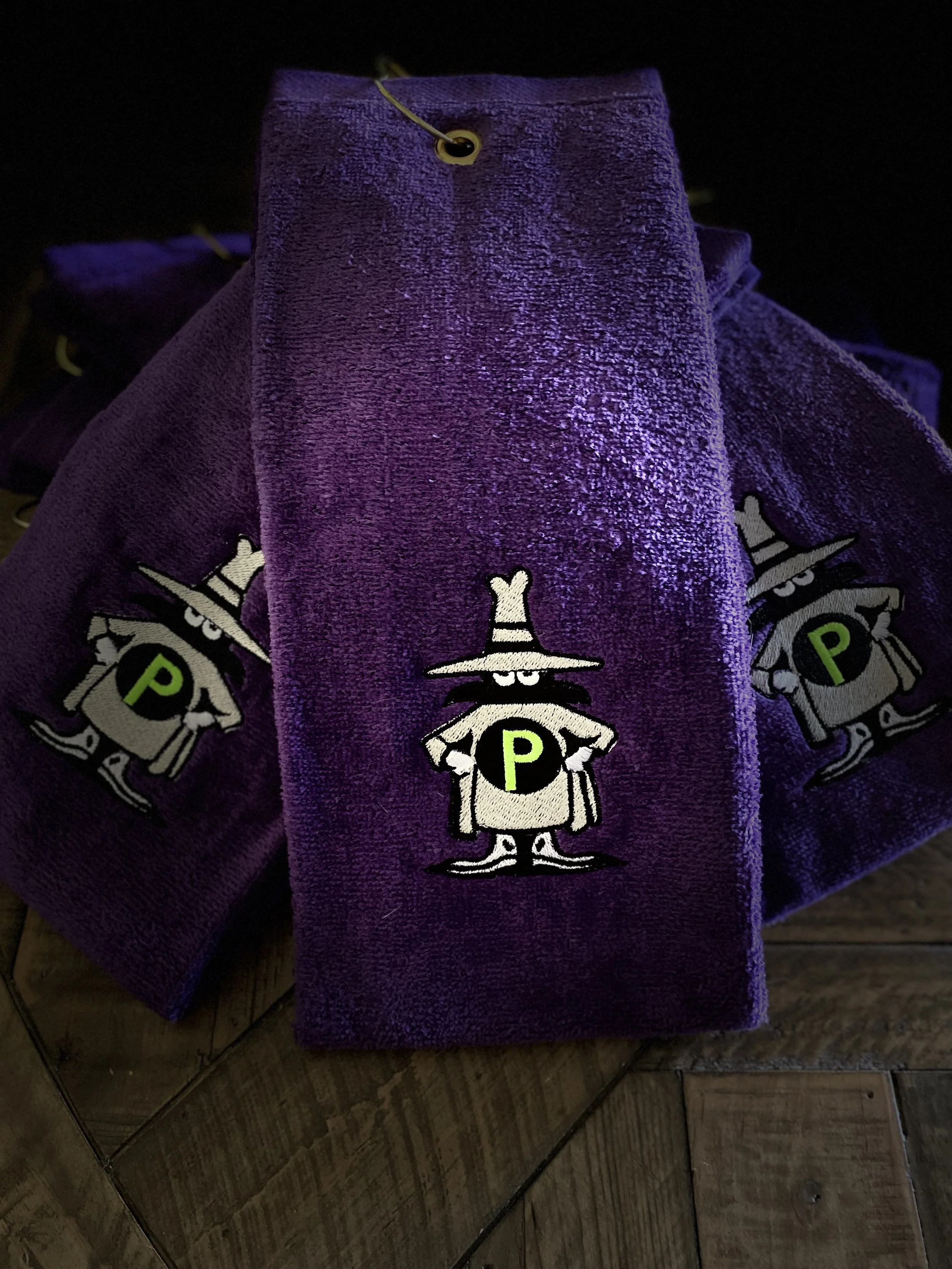 A purple towel with a cartoon witch character wearing a hat and holding a broom. The towel is hanging on a hook with a black and purple object in the background.