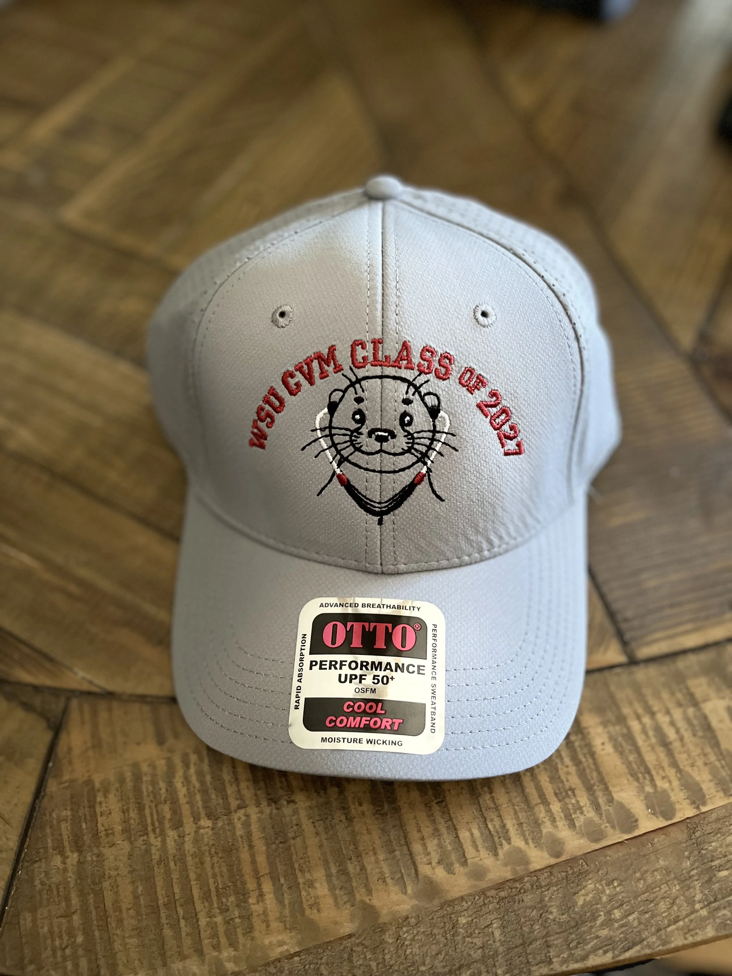 A beige baseball cap featuring a smiling lion face with the text 'WSSD CUBS GYM CLASS OF 2027' embroidered in red. The cap also has a sticker on the brim displaying its features like UPF 50+ sun protection, moisture-wicking, and cool comfort, placed on a wooden surface.