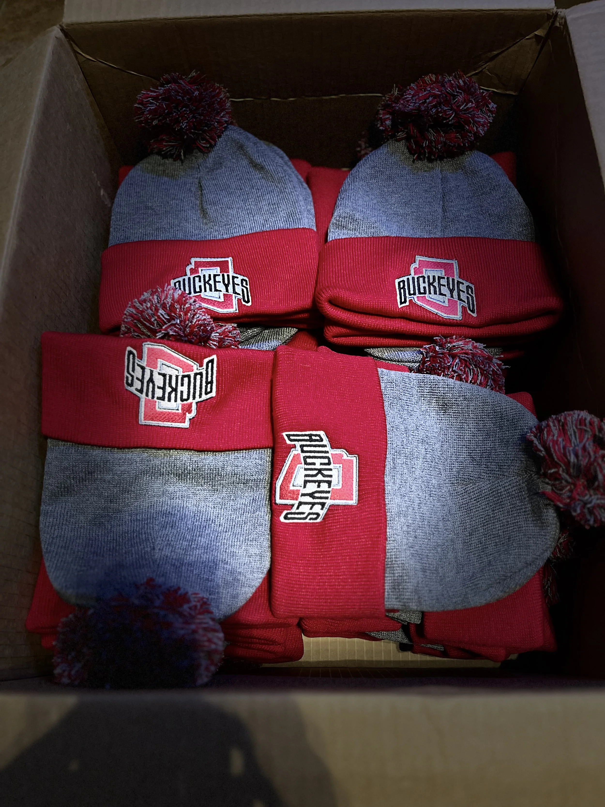 A box containing multiple gray and red knit winter hats with pom-poms on top, each hat displaying a 'Buckeyes' logo.