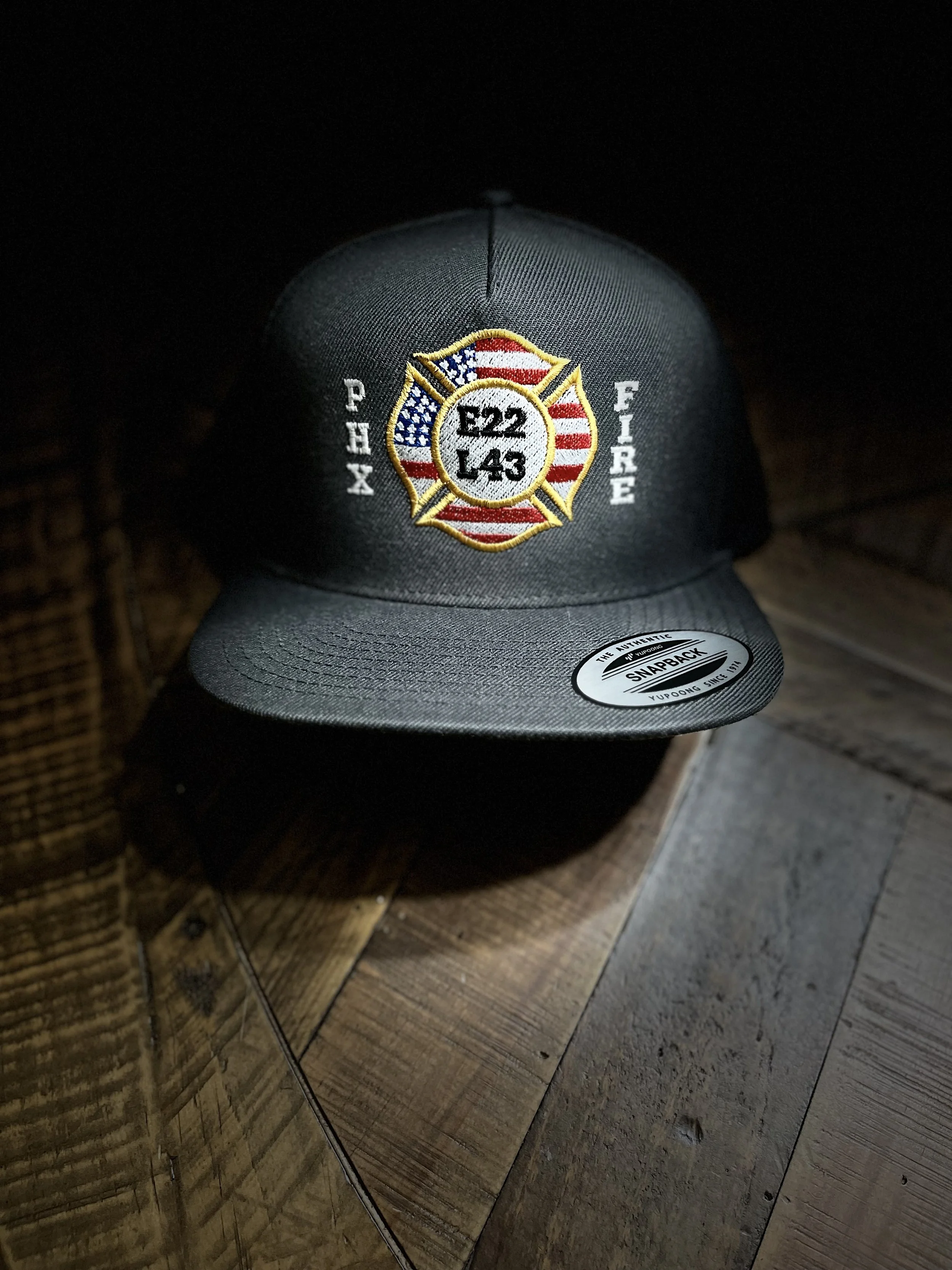 Dark gray baseball cap with an embroidered firefighter emblem on the front, featuring the American flag and the text 'E22 L43' in the center, placed on a wooden surface.