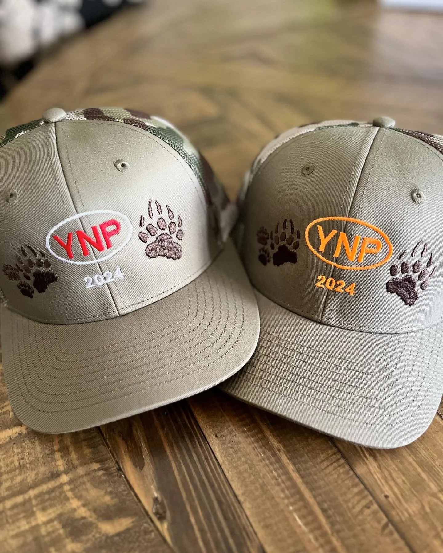 Two beige baseball caps with embroidered logos reading 'YNP 2024' and paw prints, placed on a wooden surface.