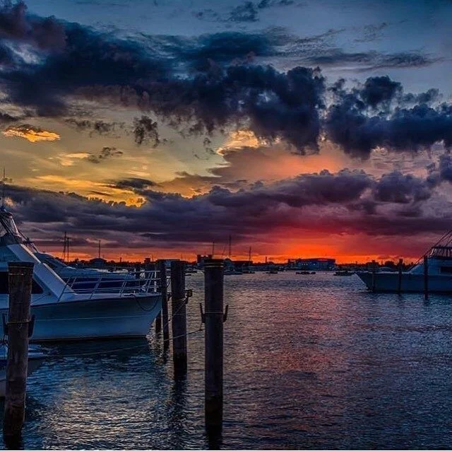 Boats docked at a marina during a colorful sunset with dark clouds overhead.