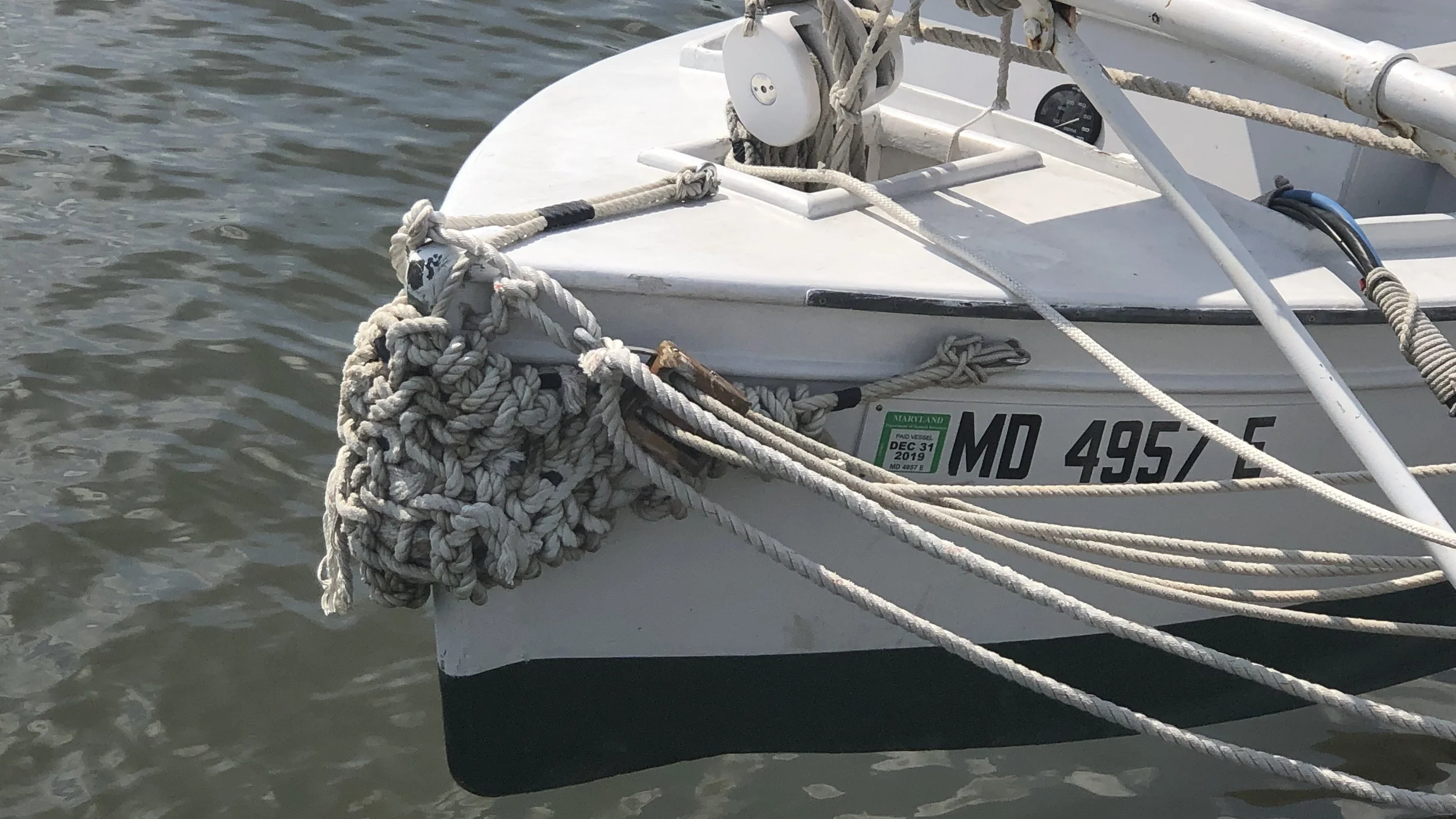 Close-up of the bow of a white sailboat tied to a dock, with numerous coiled ropes and rigging, and a Maryland registration sticker on the side.