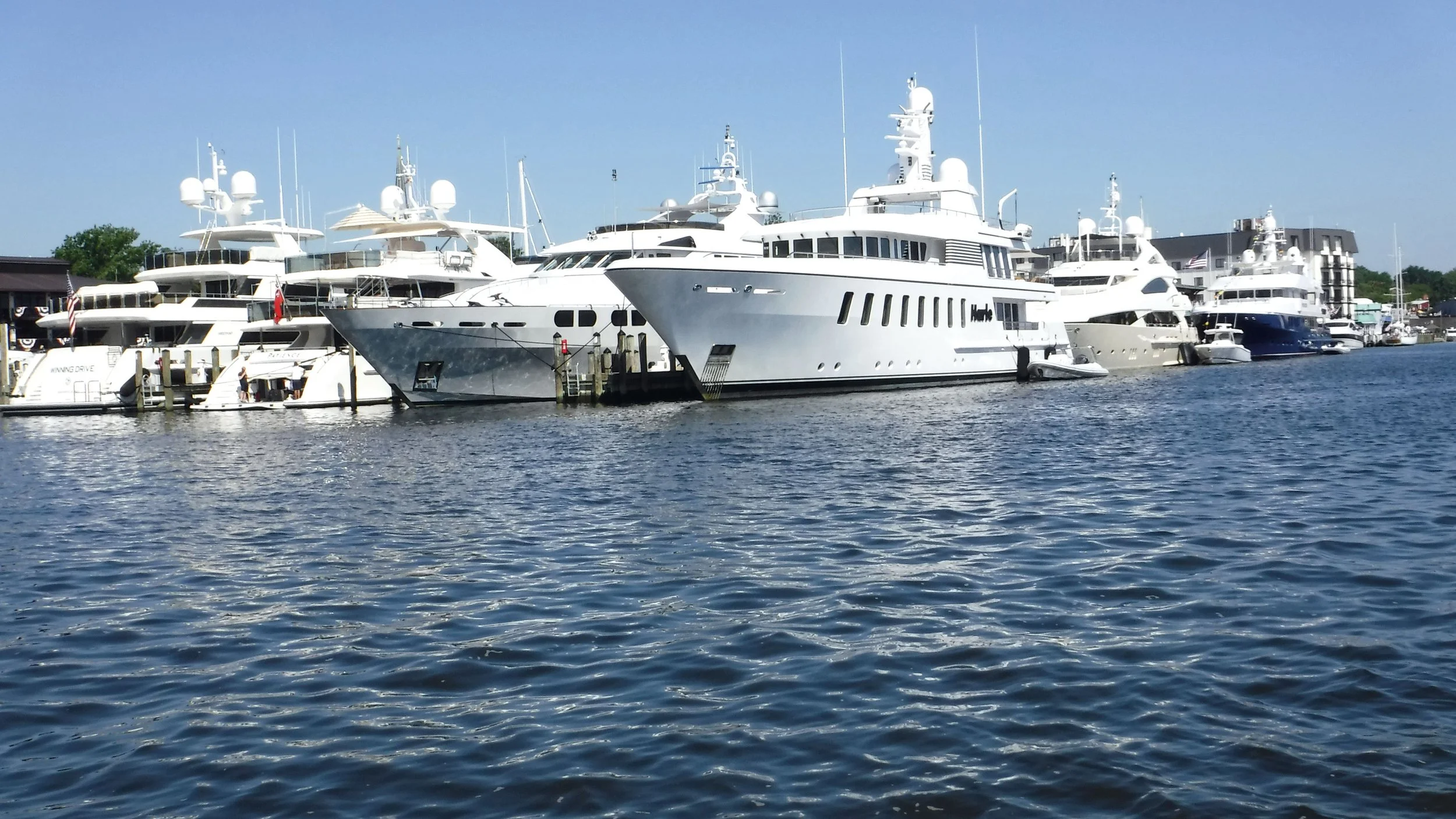 Several large yachts docked at a marina on a sunny day, with calm water and a clear blue sky.