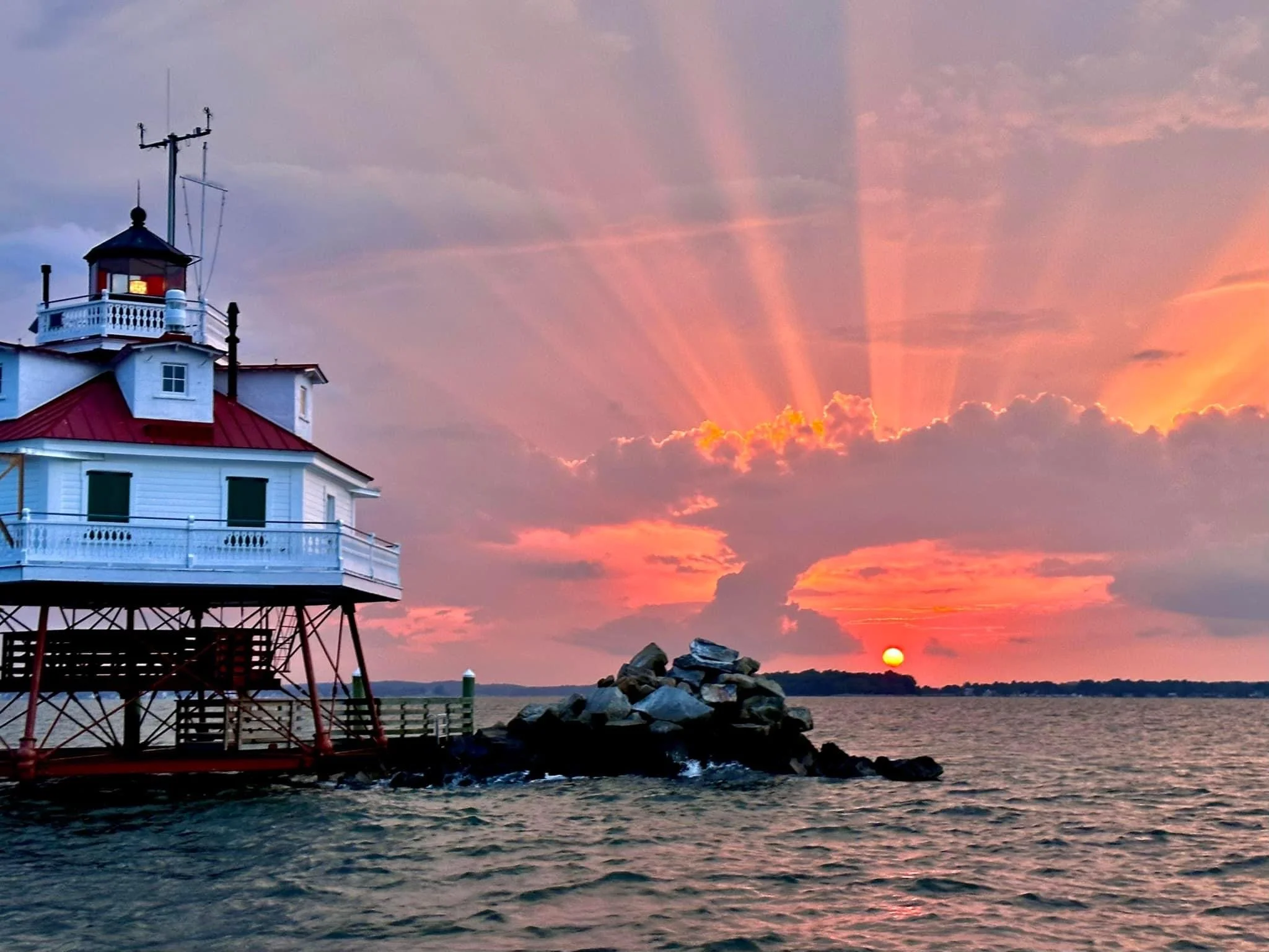 A lighthouse on stilts at sunset with a colorful sky and rays of sunlight emanating from the horizon over a body of water.
