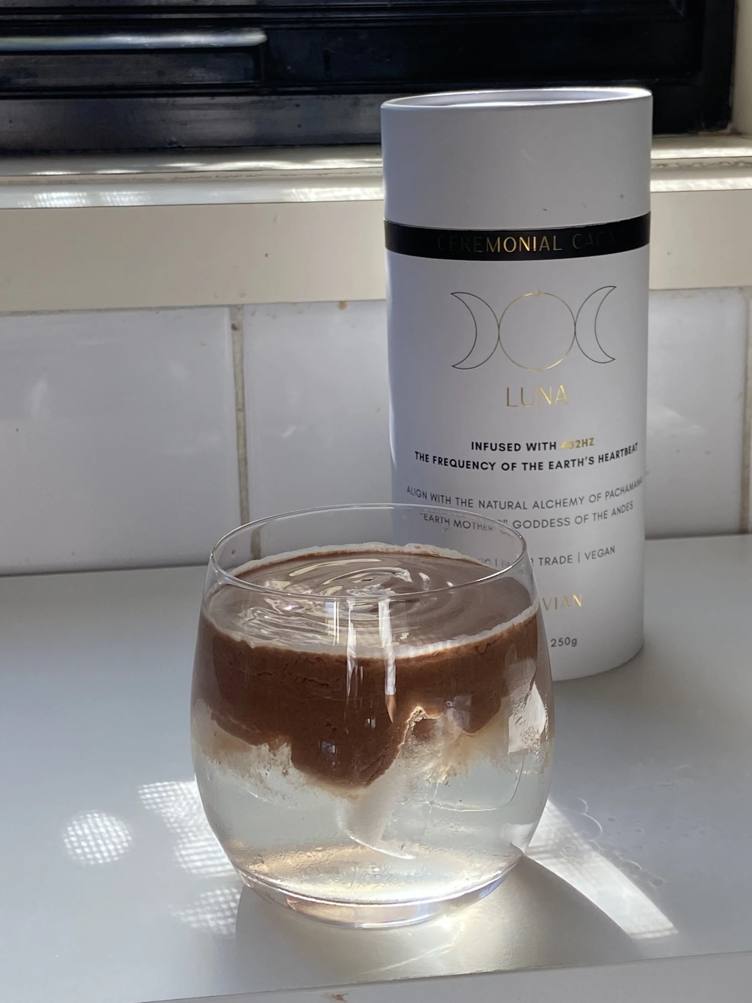 A glass of layered chocolate and vanilla dessert with a foamy top, placed on a white surface in front of a window. In the background, there is a white canister labeled "CEREMONIAL CACE LUNA".