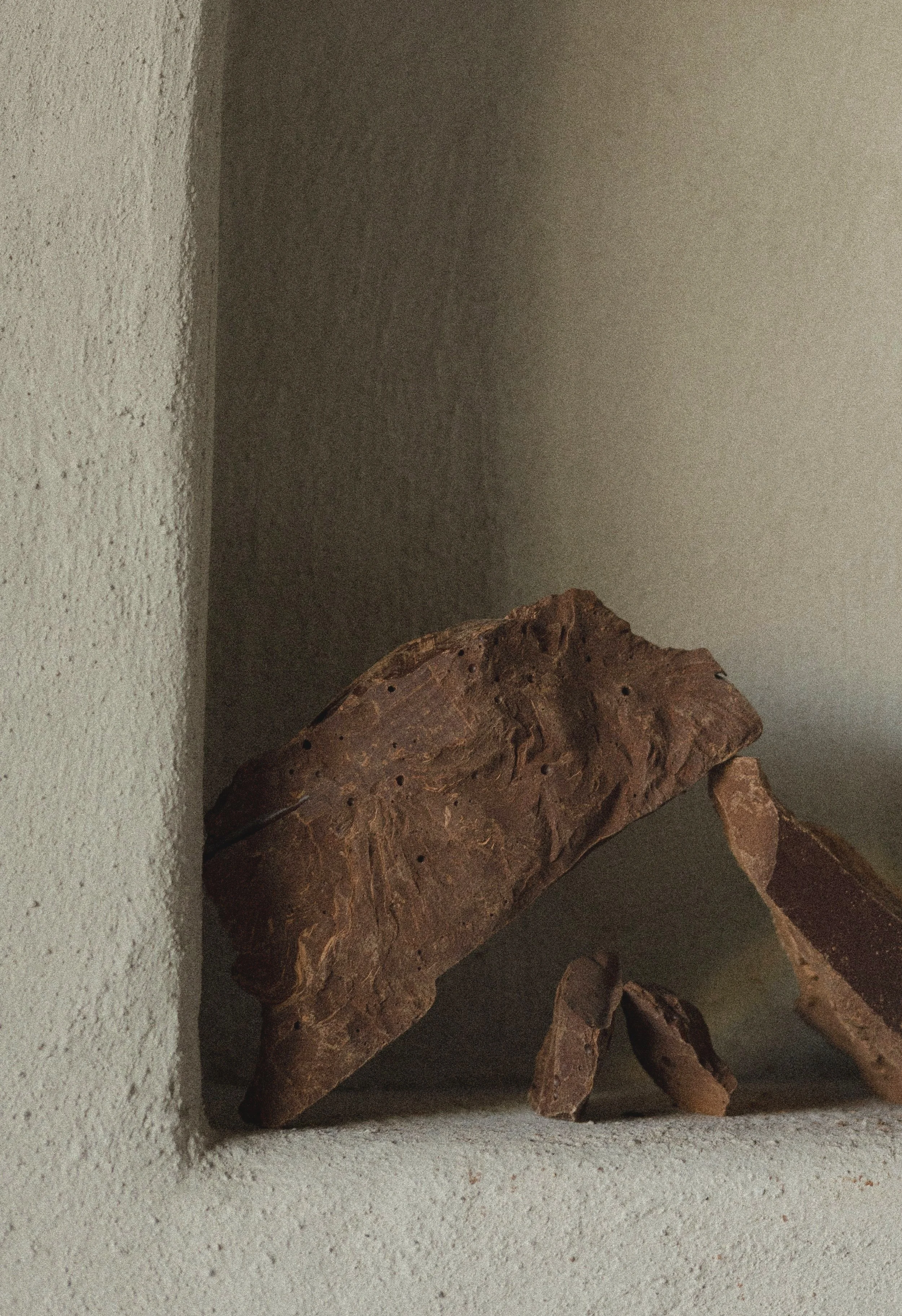 A wooden sculpture resembling a humanoid figure, placed on a white textured surface against a plain beige wall.