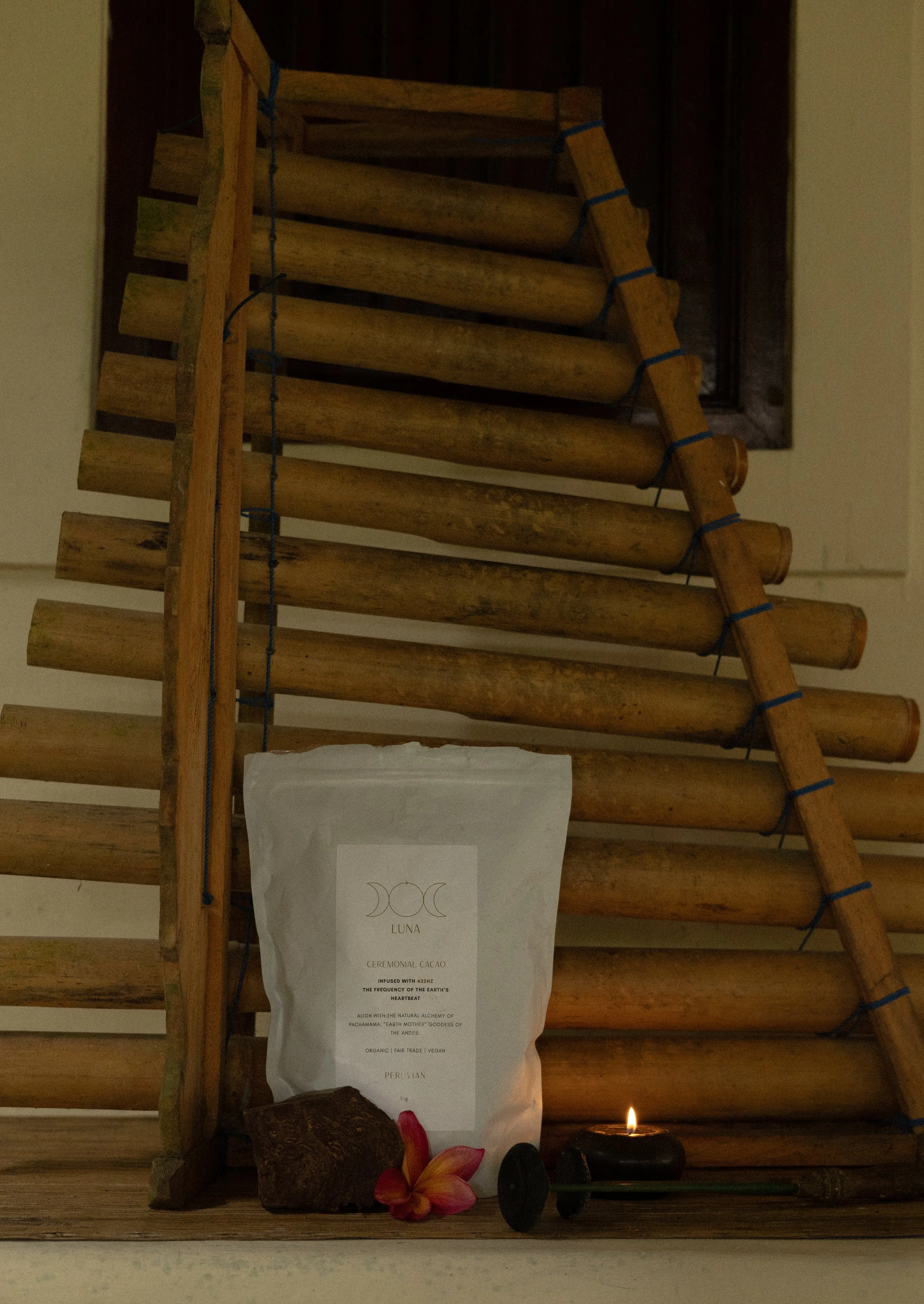 A Zen-style altar with a wooden ladder, a white paper bag with the text "LUNA CEREMONIAL CACAO," a flower, stones, and a lit candle.
