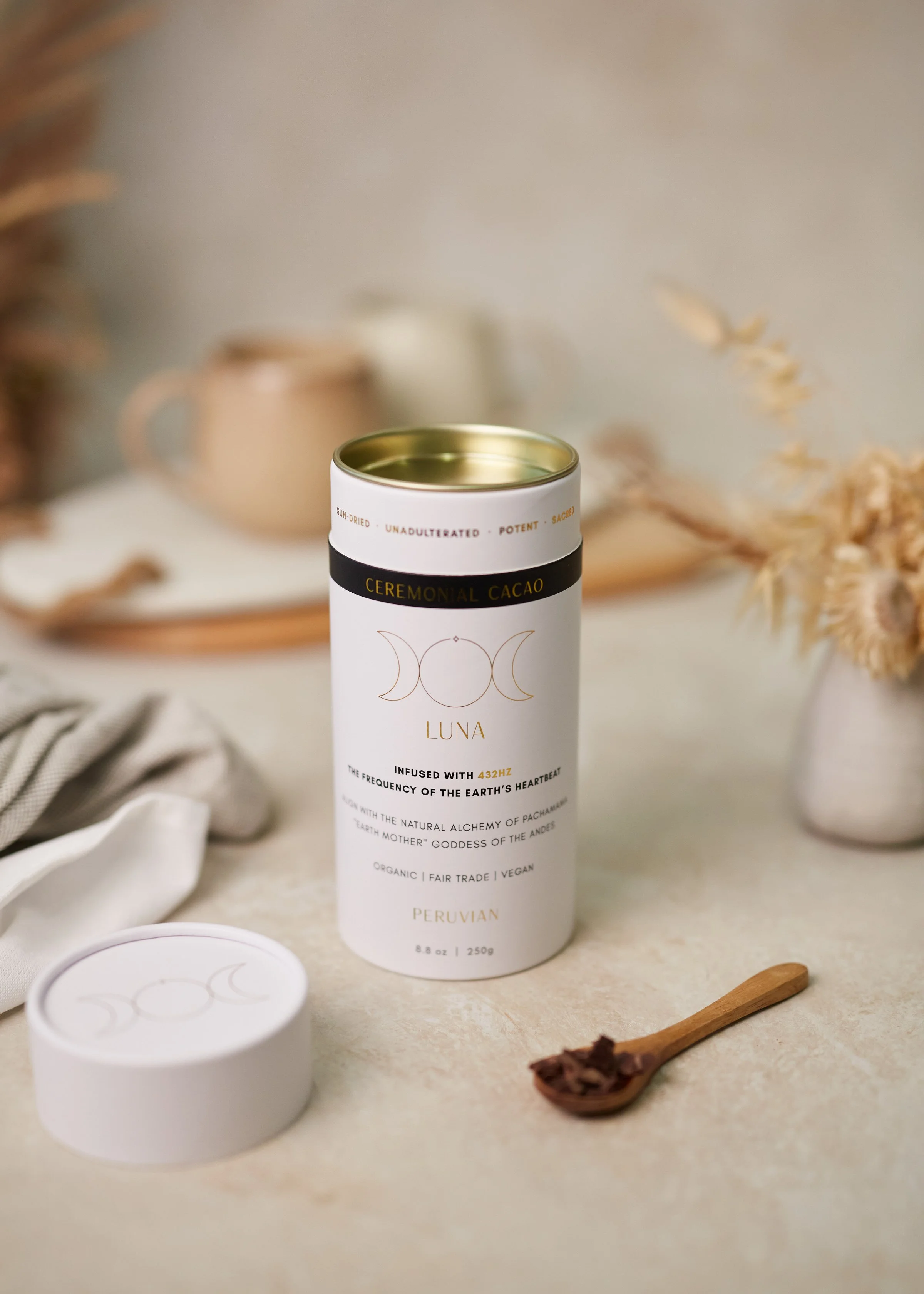 A tall white cylindrical container of ceremonial cacao with a gold lid, placed on a beige surface with a wooden spoon filled with cacao paste, and blurred background of dried plants, a teapot, and cloth.