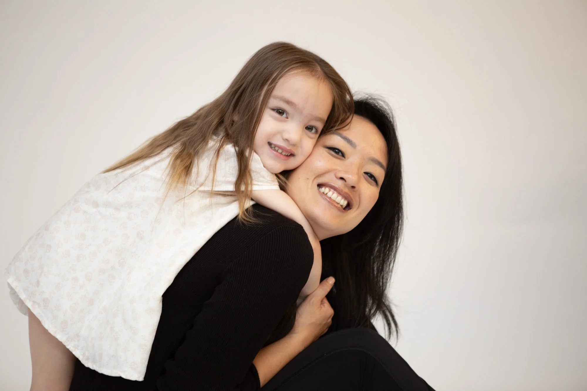 A woman with long dark hair smiling while carrying a young girl with long light brown hair on her back, both posing against a plain white background.