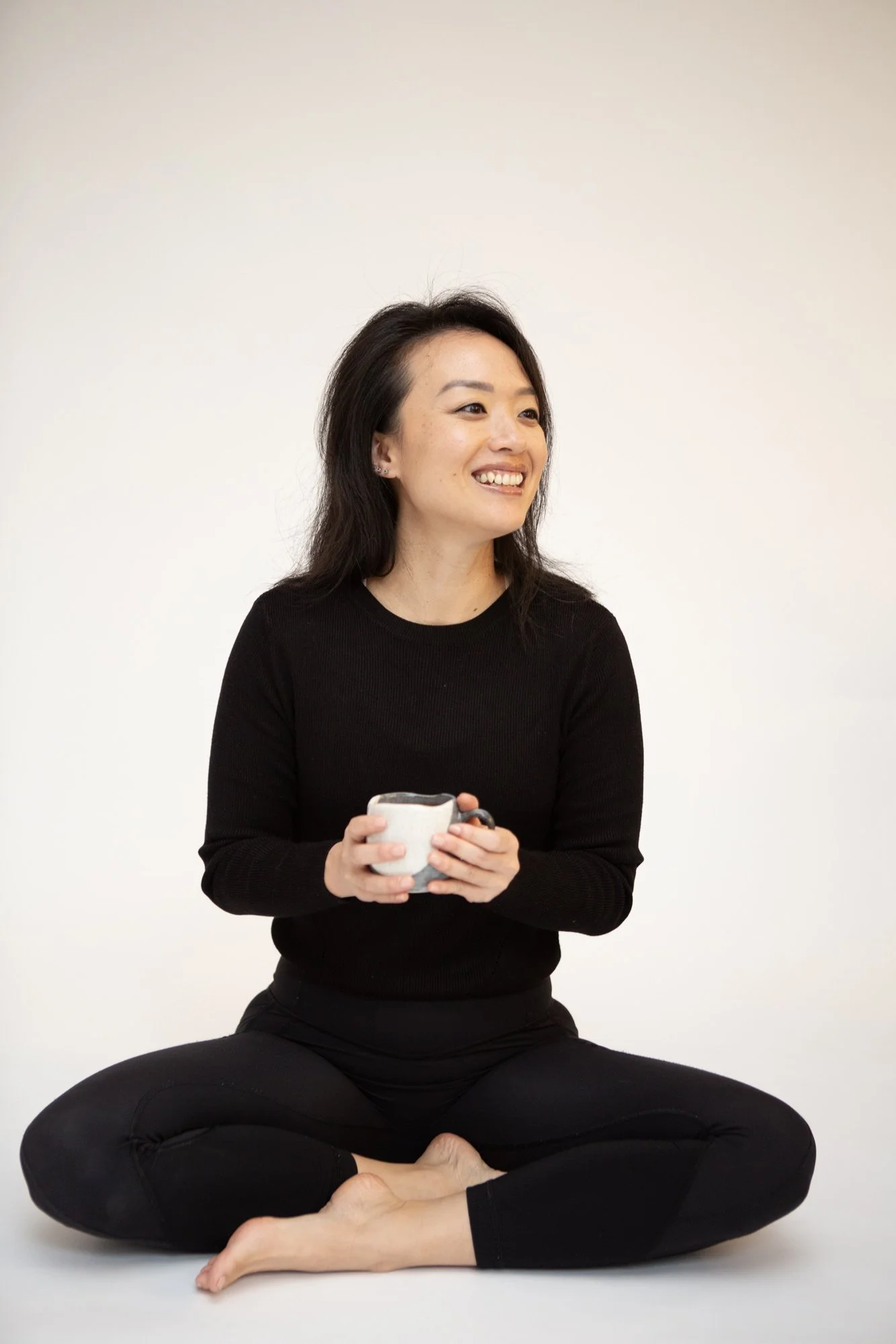 A woman with dark hair sitting cross-legged in a meditative pose on the floor against a plain white background. She is smiling, holding a ceramic mug in both hands, wearing a black long-sleeve shirt and black leggings.